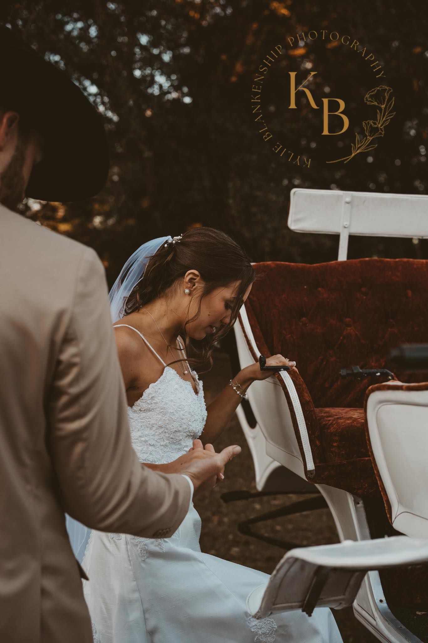 Bride in lace gown stepping into a vintage car assisted by groom in tan suit at outdoor wedding.