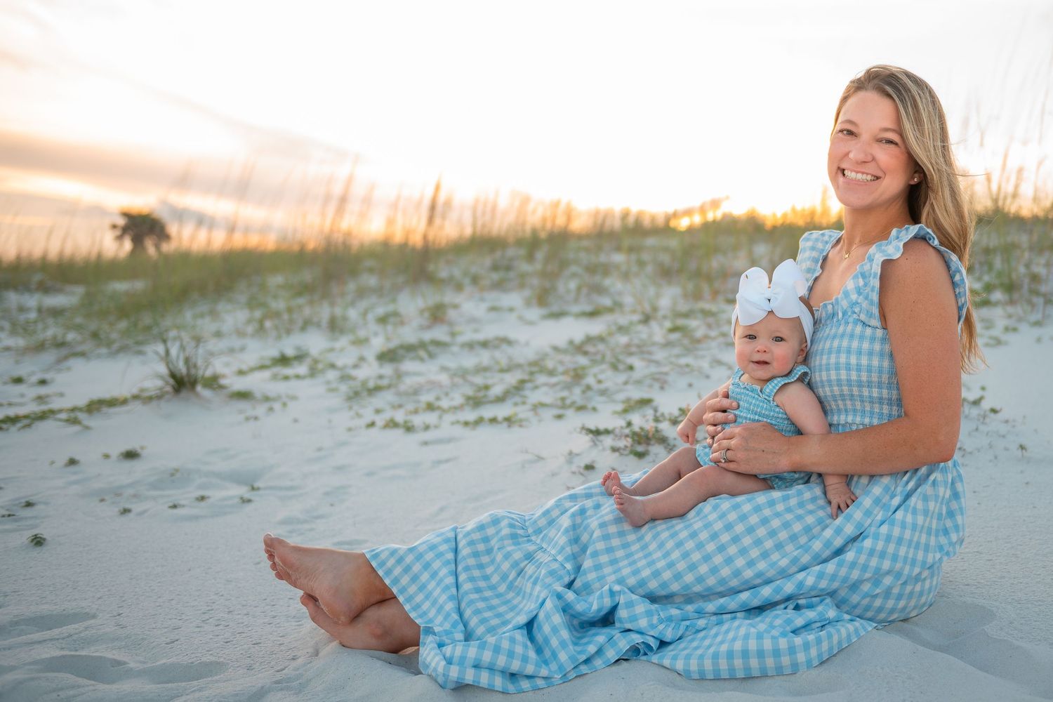 Smiling mother in blue gingham dress sitting on beach sand holding her baby girl.