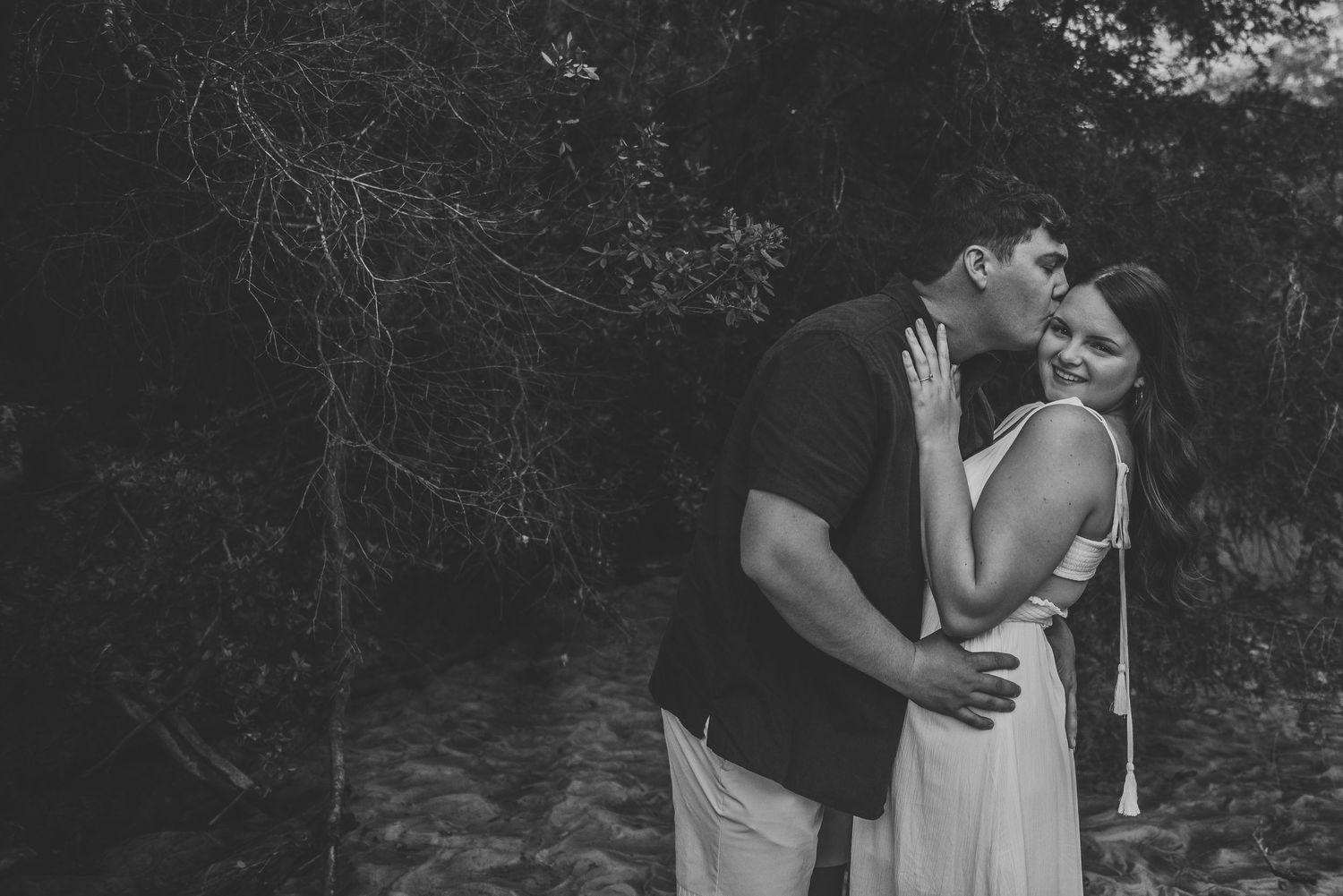 Black and white romantic photo of couple embracing tenderly in a wooded outdoor setting.