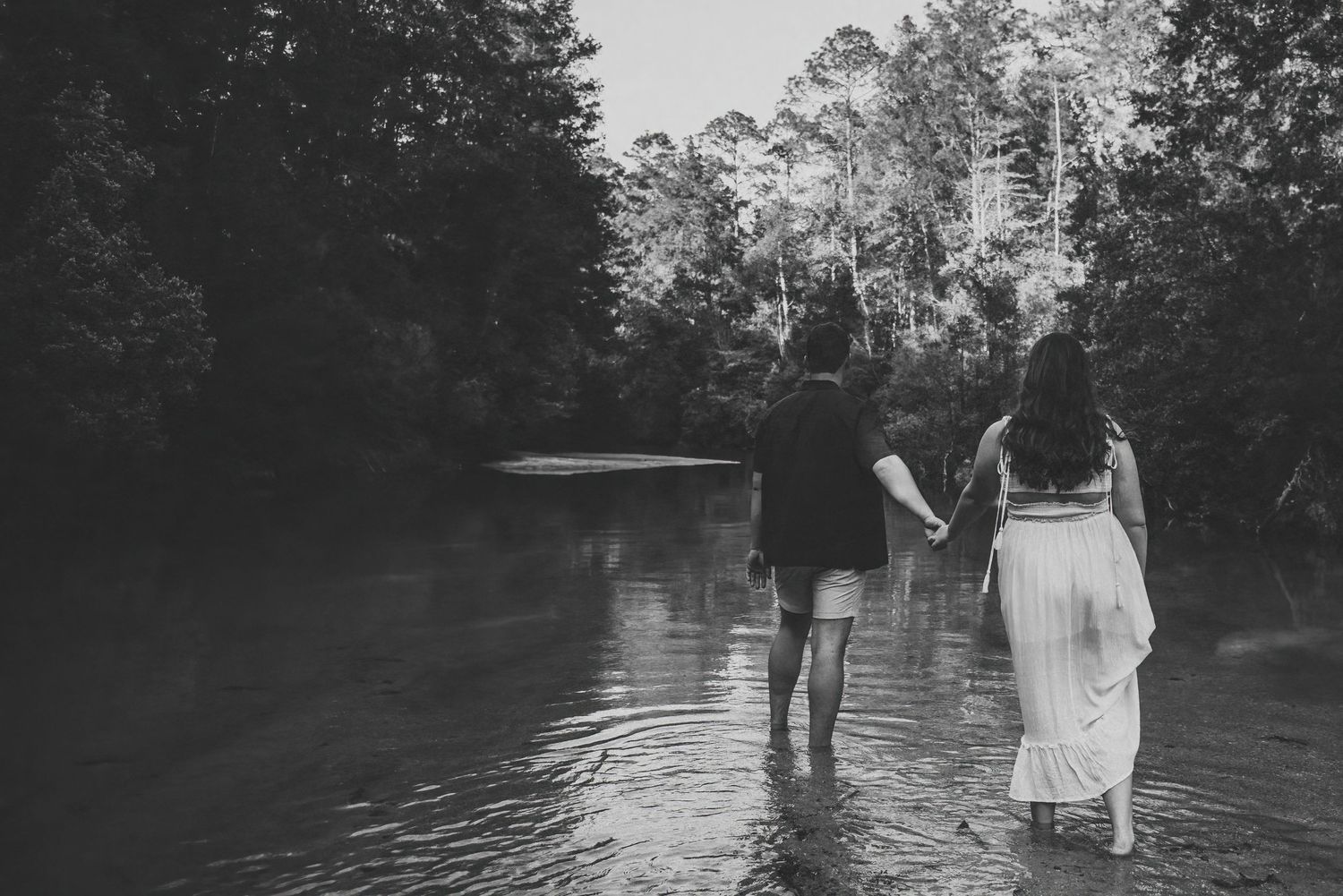 Black and white photo of couple holding hands wading through a serene forest river.
