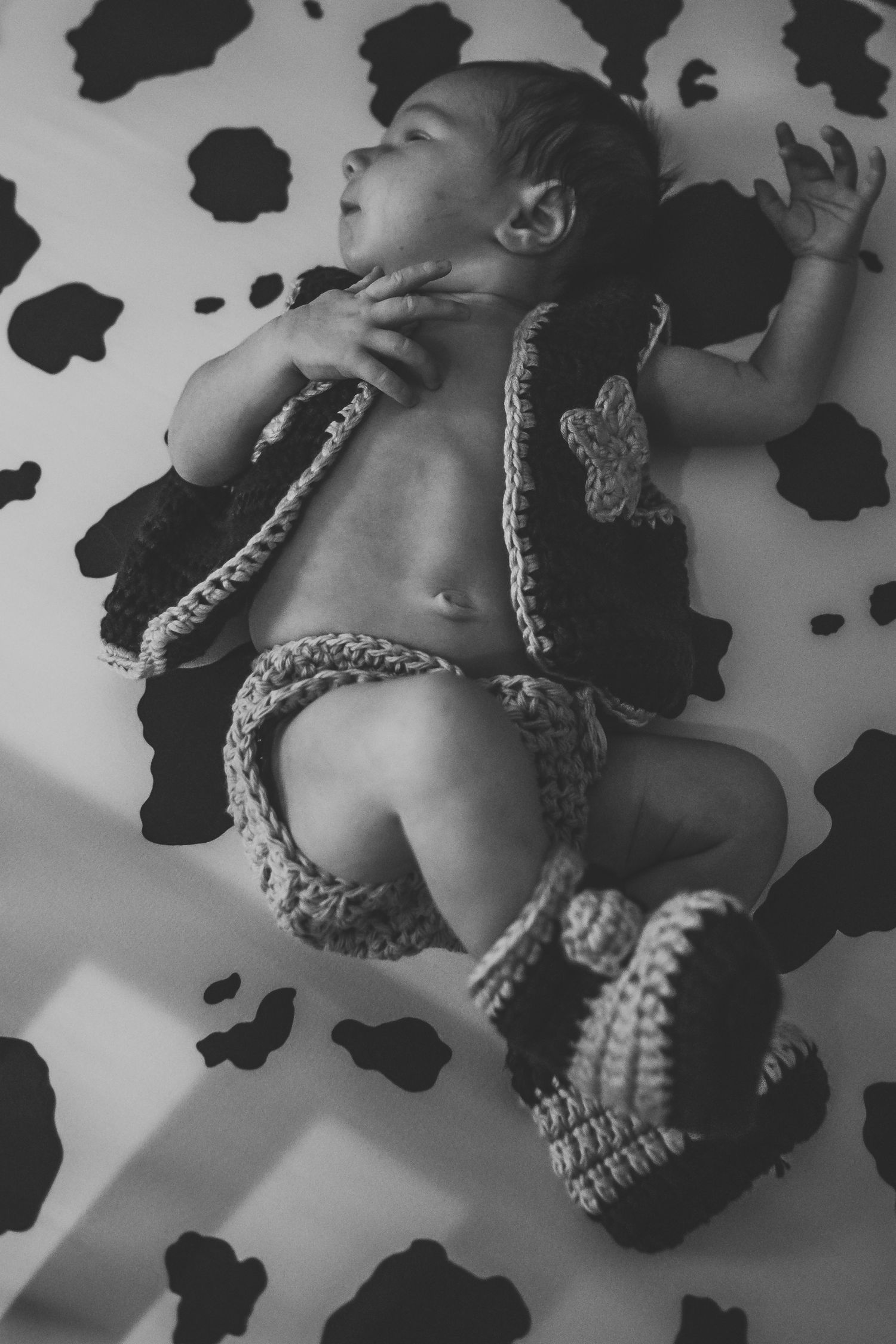 Black and white photo of sleeping newborn in crocheted cowboy outfit on cow print sheet.