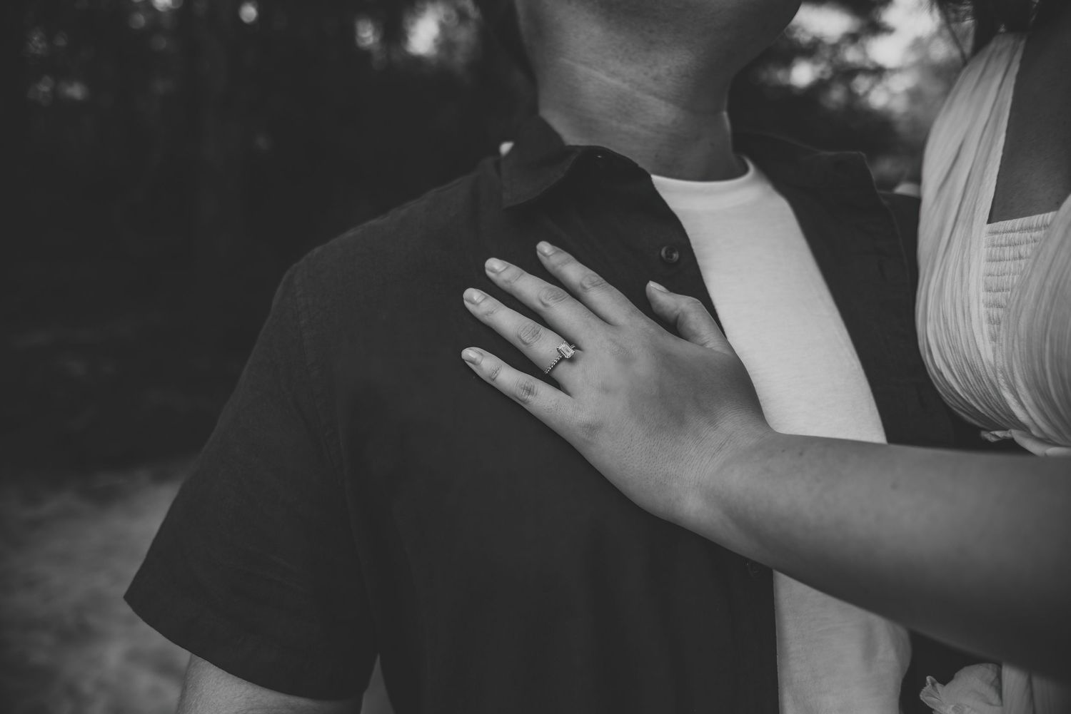 Black and white close-up of a woman's engagement ring as she places her hand on her partner's chest.