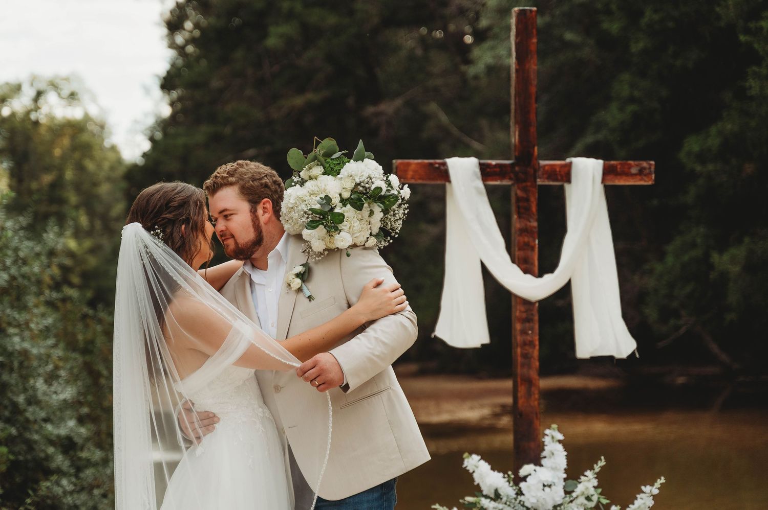 Bride and groom kissing at outdoor wedding ceremony beside a rustic wooden cross with white draping.