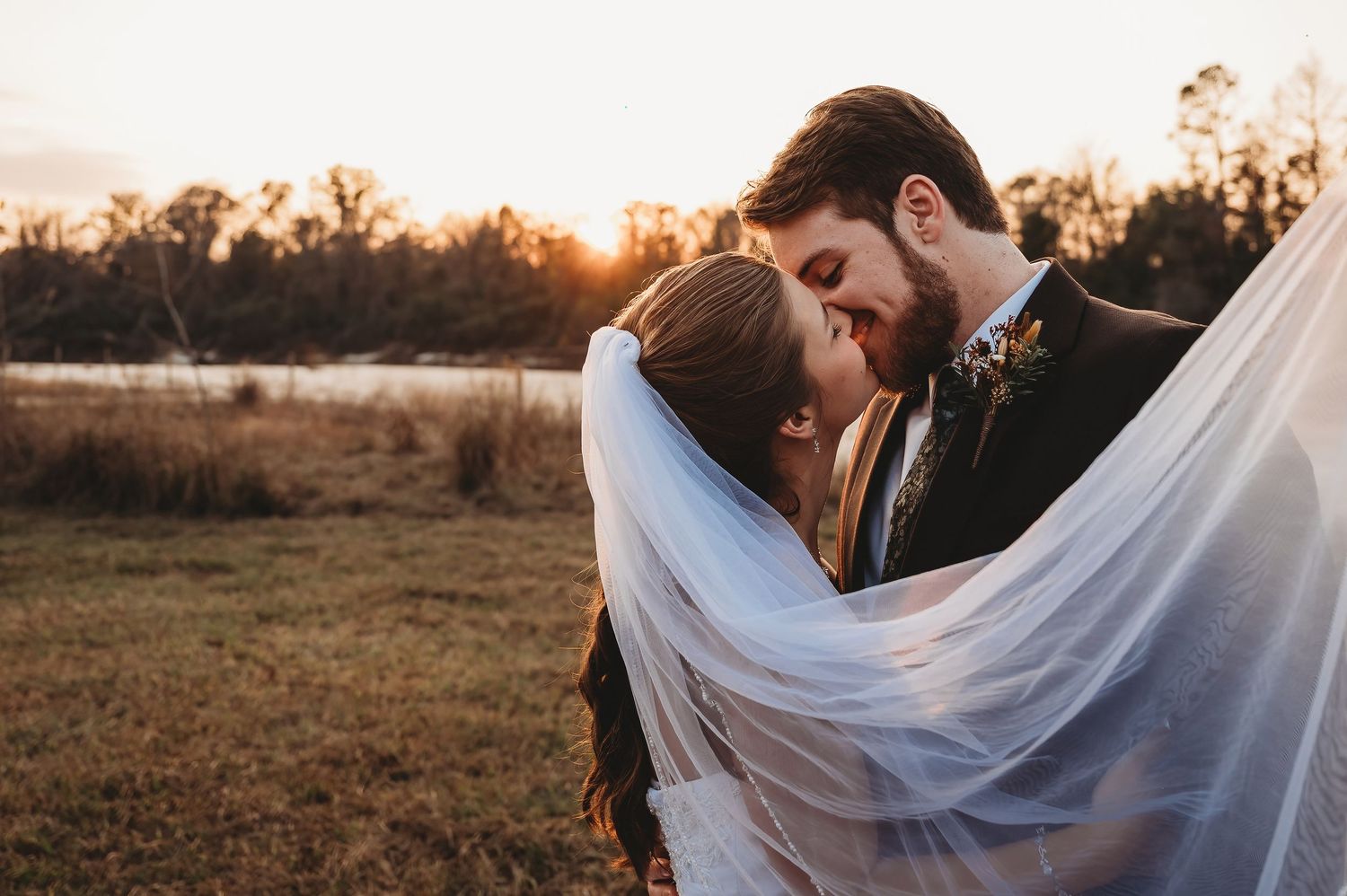 Couple nearly kissing under a bridal veil with warm sunset light glowing behind them outdoors.