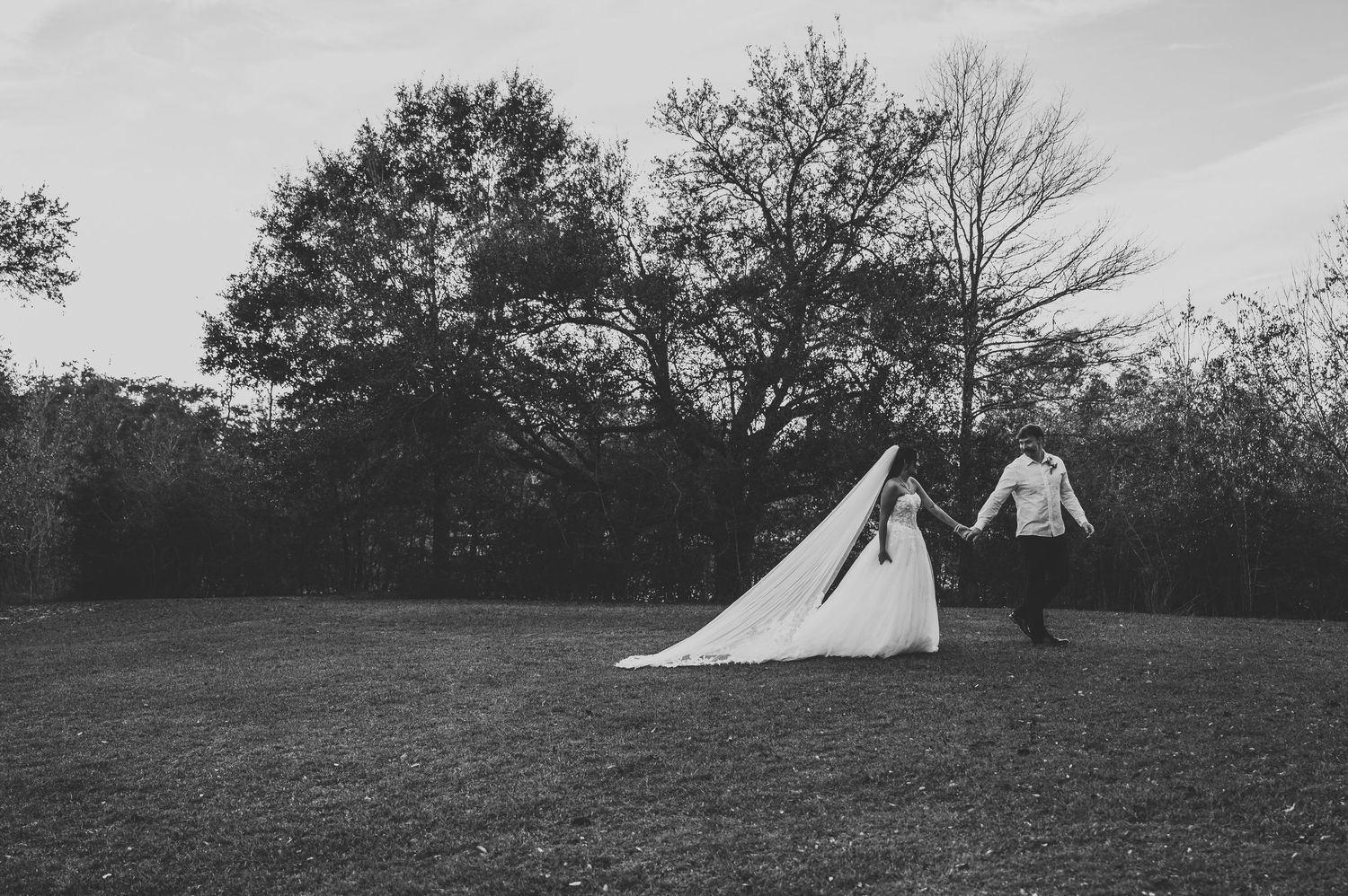 Black and white photo of bride and groom holding hands walking across a lush green lawn with trees in the background.
