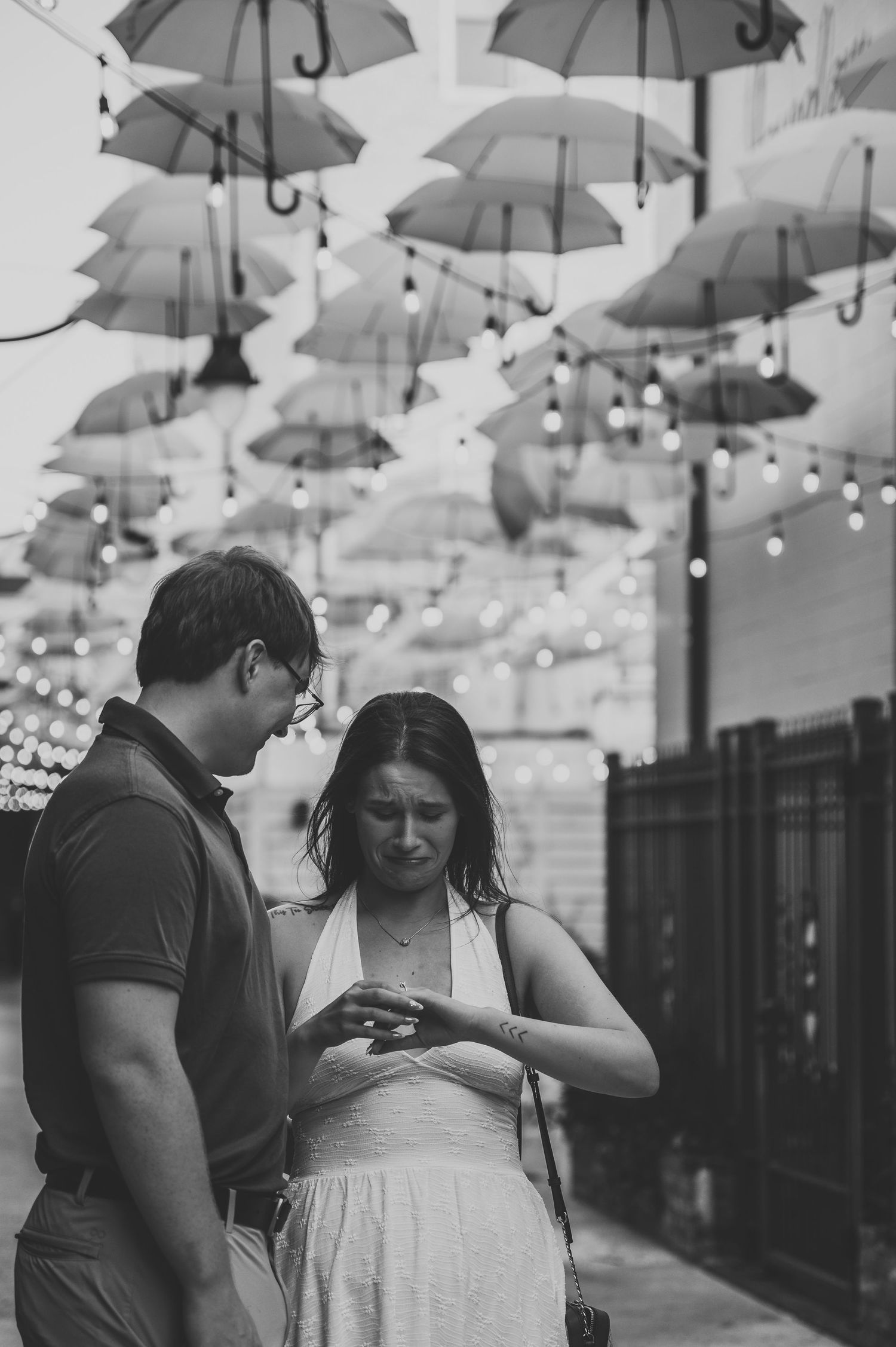 Black and white photo of a couple looking at an engagement ring under hanging umbrellas and string lights.