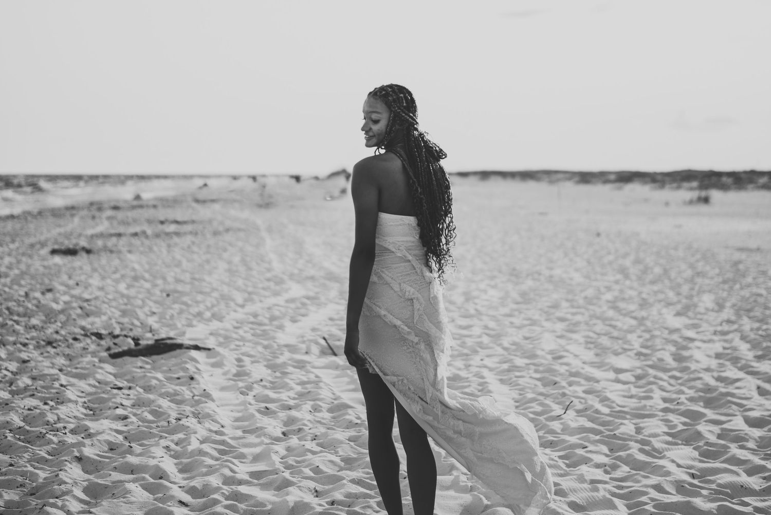 Black and white photo of young woman in strapless white dress standing on sandy beach looking away.