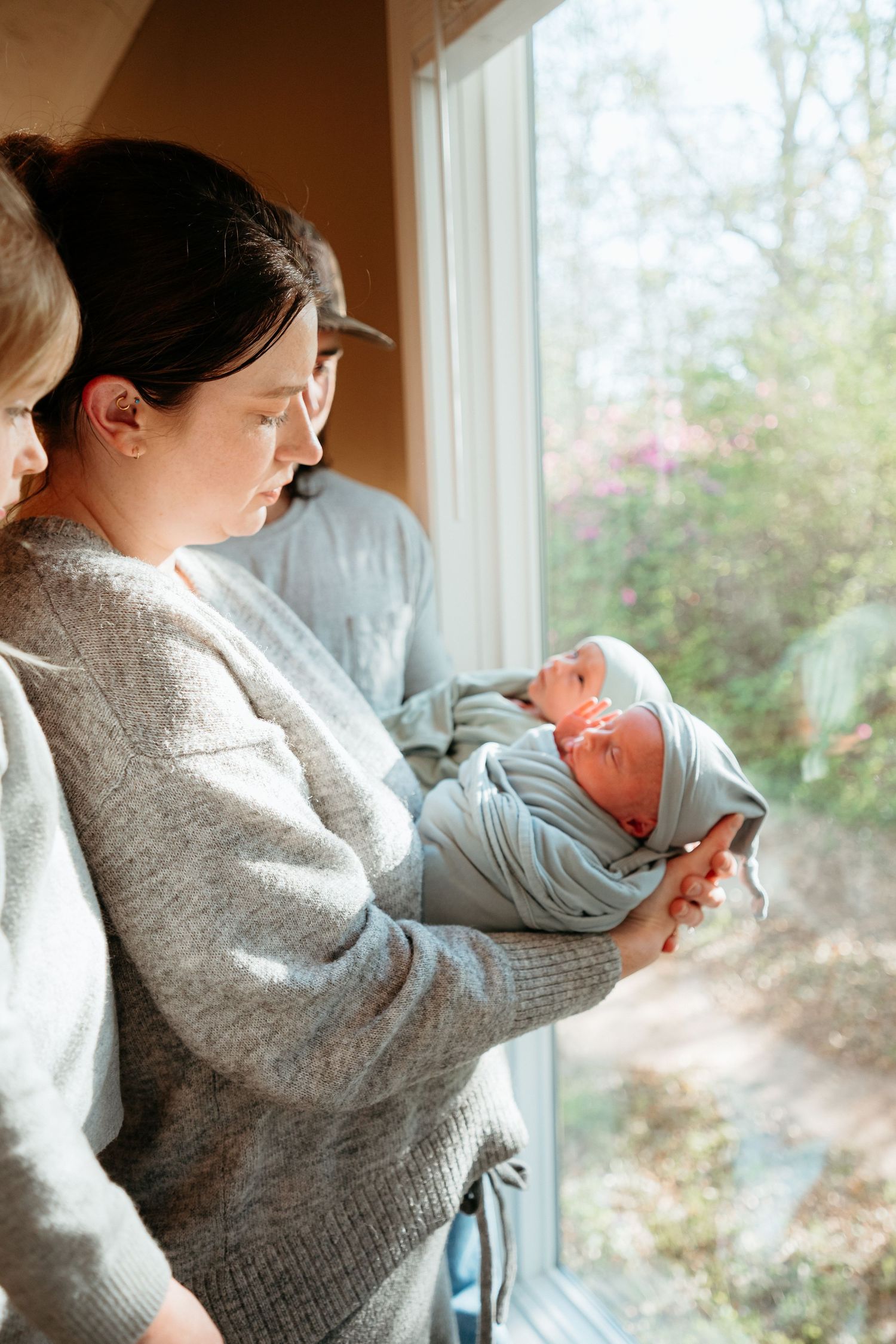 Mother holds swaddled newborn in light blue wrap near sunny window while toddler looks on.