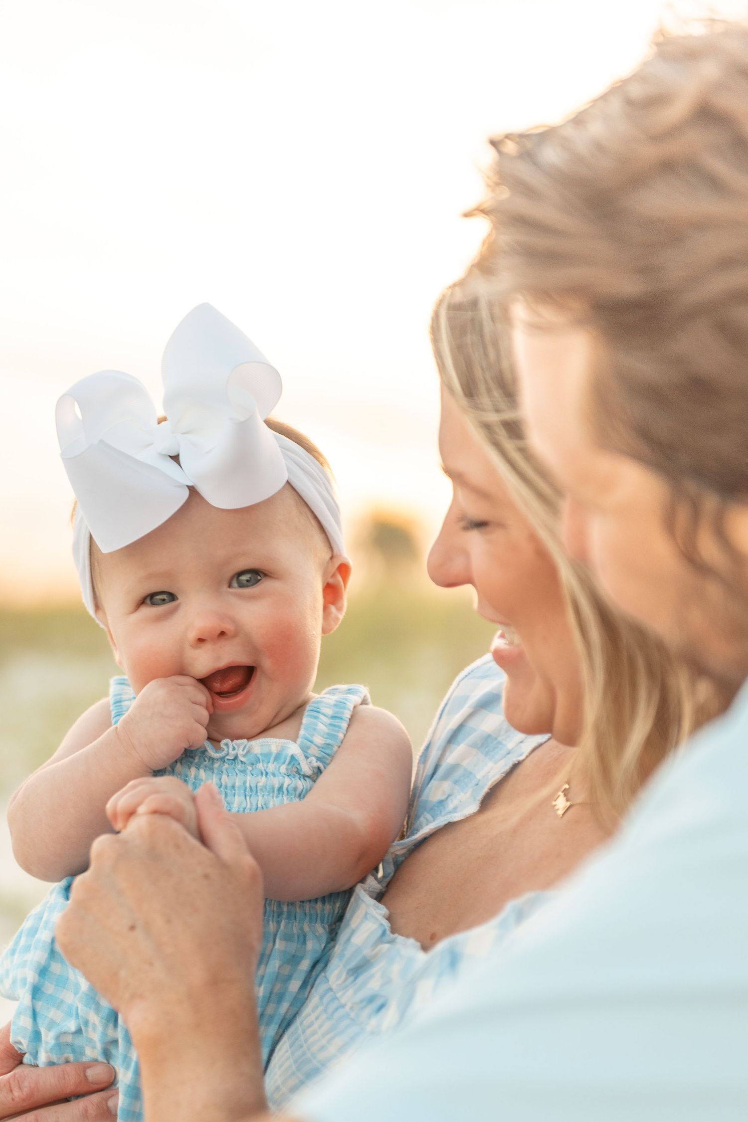Smiling baby girl wearing a white bow headband and blue dress being held by her mother at golden hour.