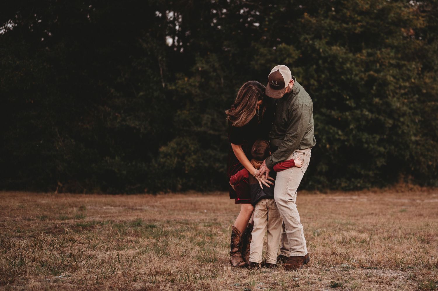 A couple and child share a tender embrace in an open field surrounded by lush green trees during golden hour.