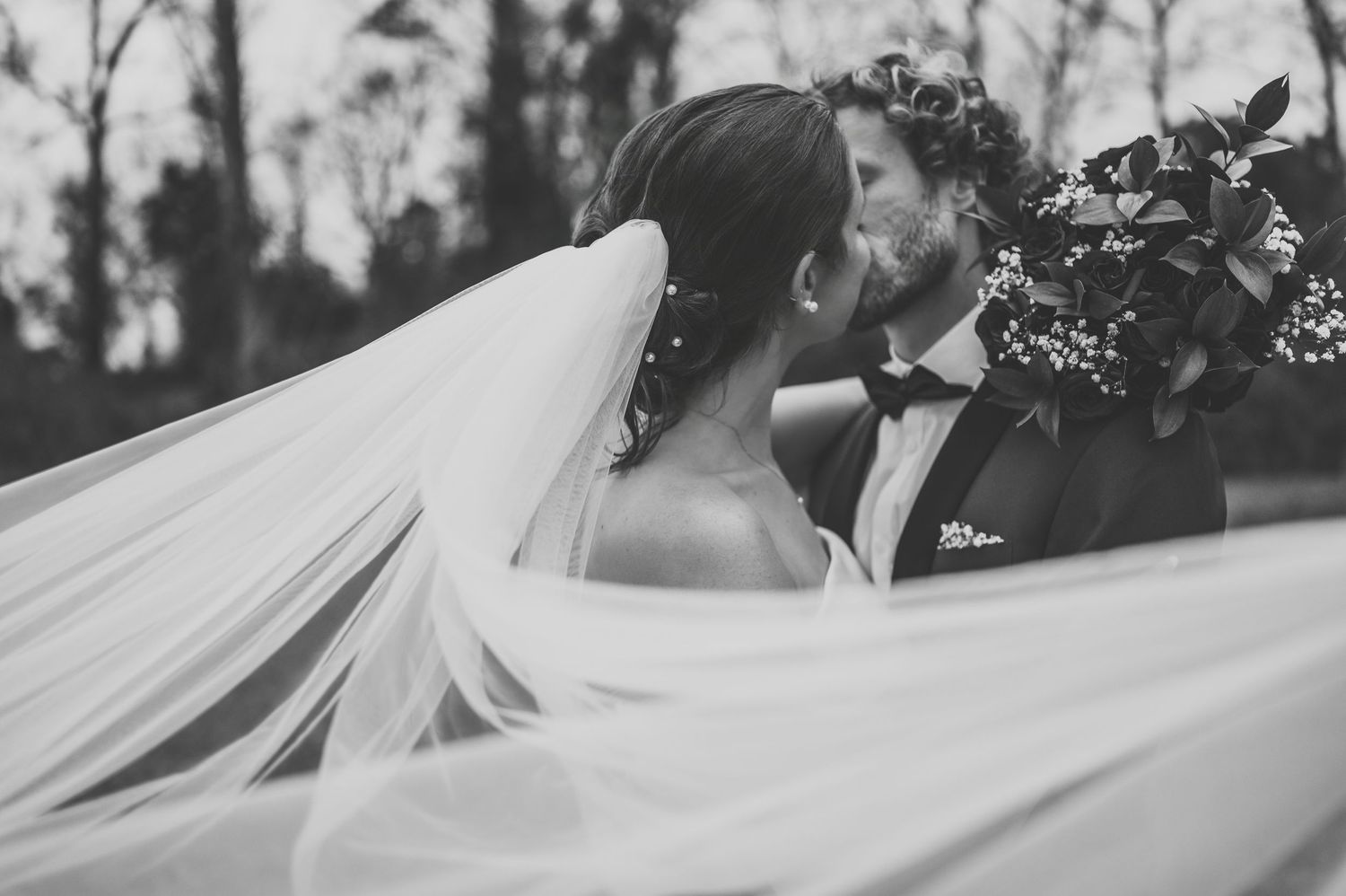 Black and white wedding photo of bride and groom kissing, veil flowing, groom in tuxedo holding floral bouquet.