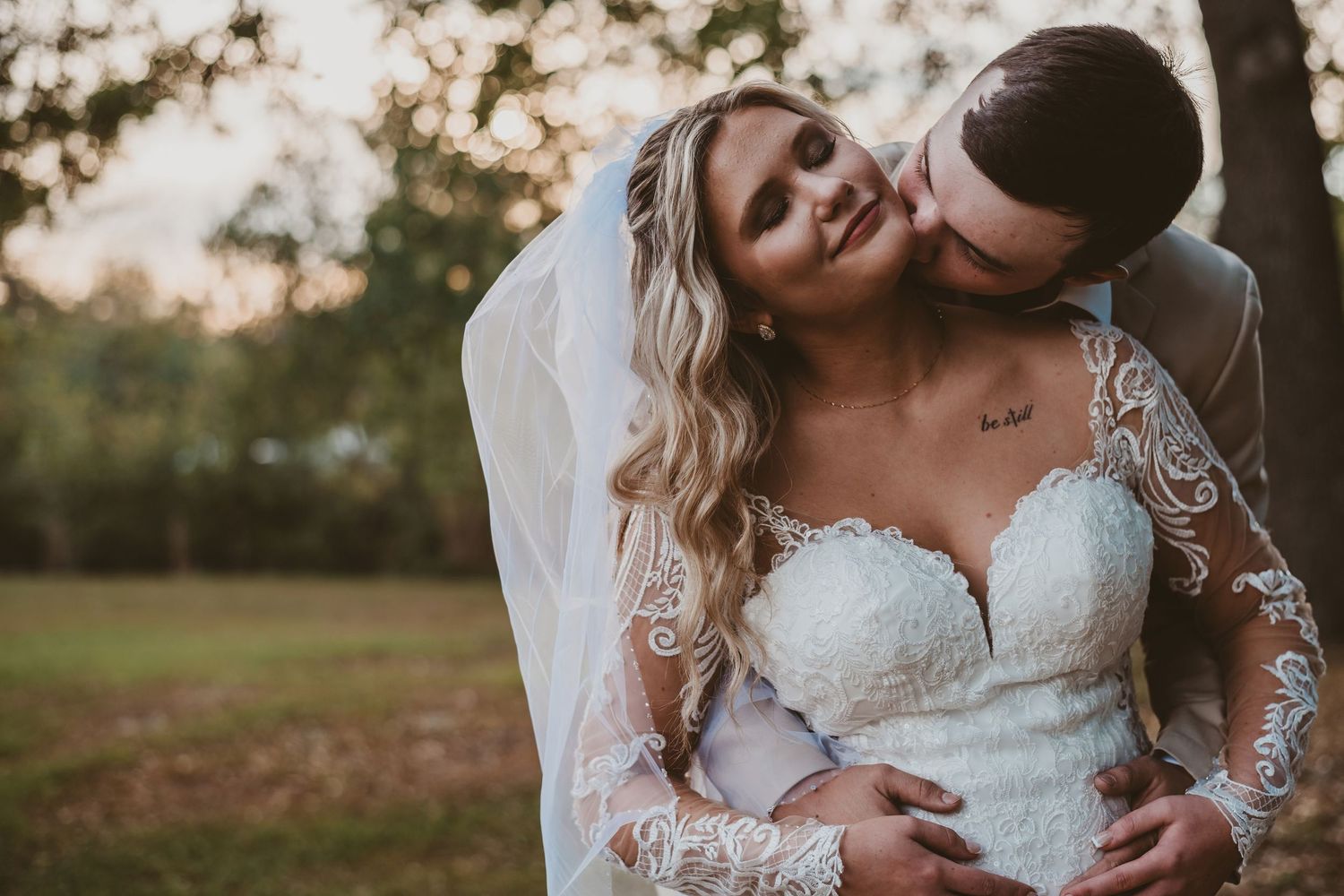 Groom tenderly kisses bride on the neck as she smiles in a lace long-sleeve wedding gown.
