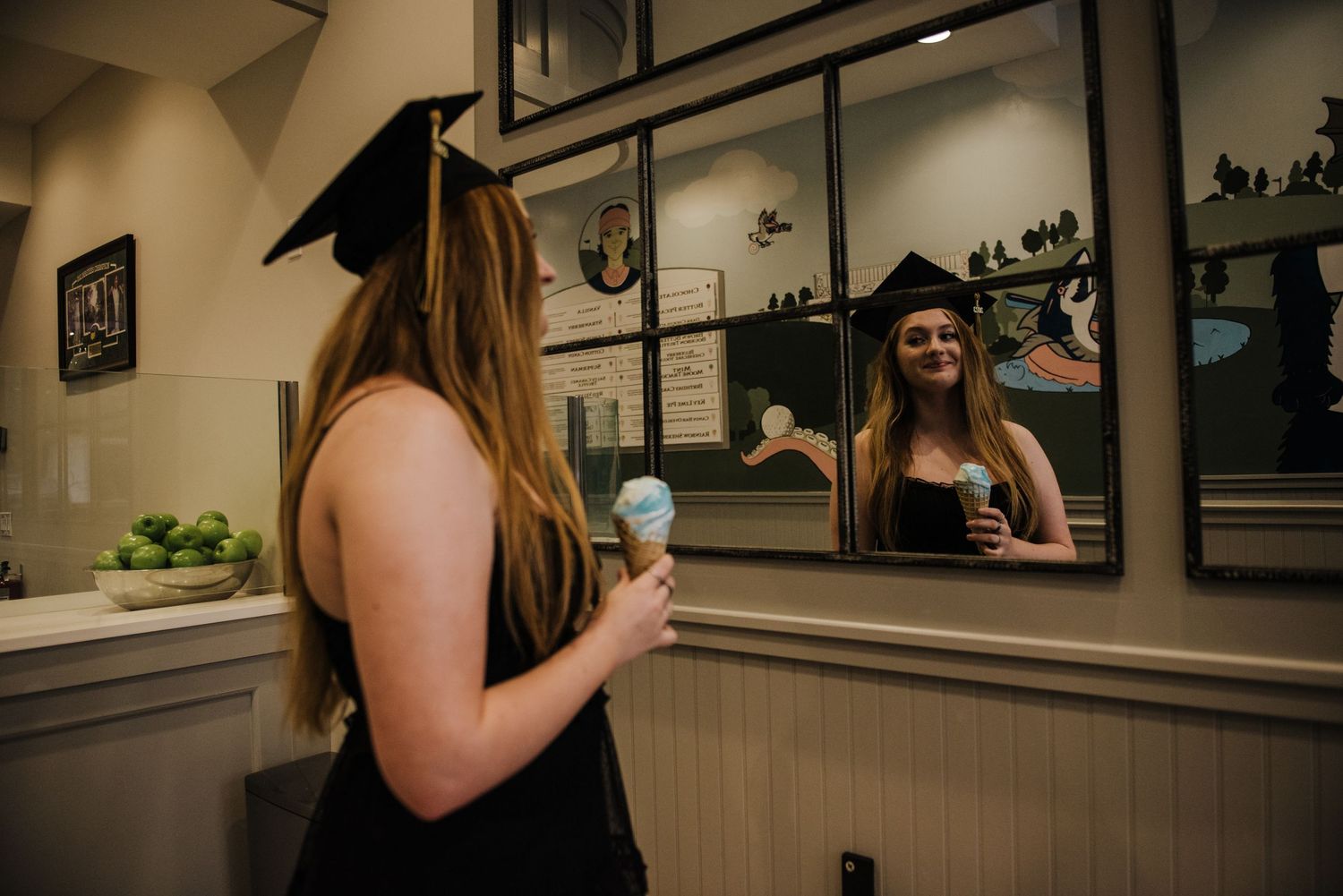 Female graduate in black cap and gown admiring her reflection in a mirror while holding a blue ice cream cone.
