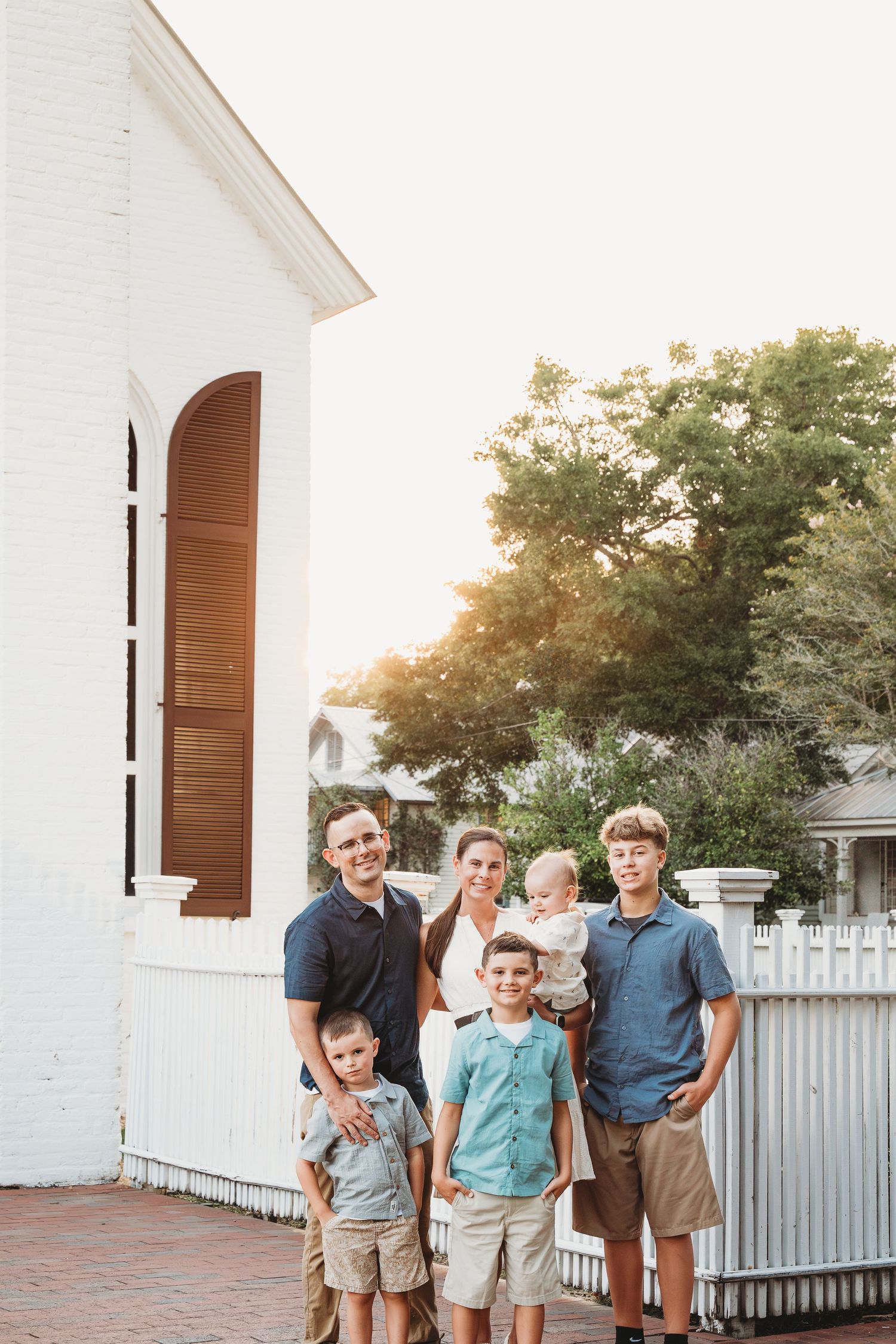 A family of six poses together outside a charming white church at golden hour.