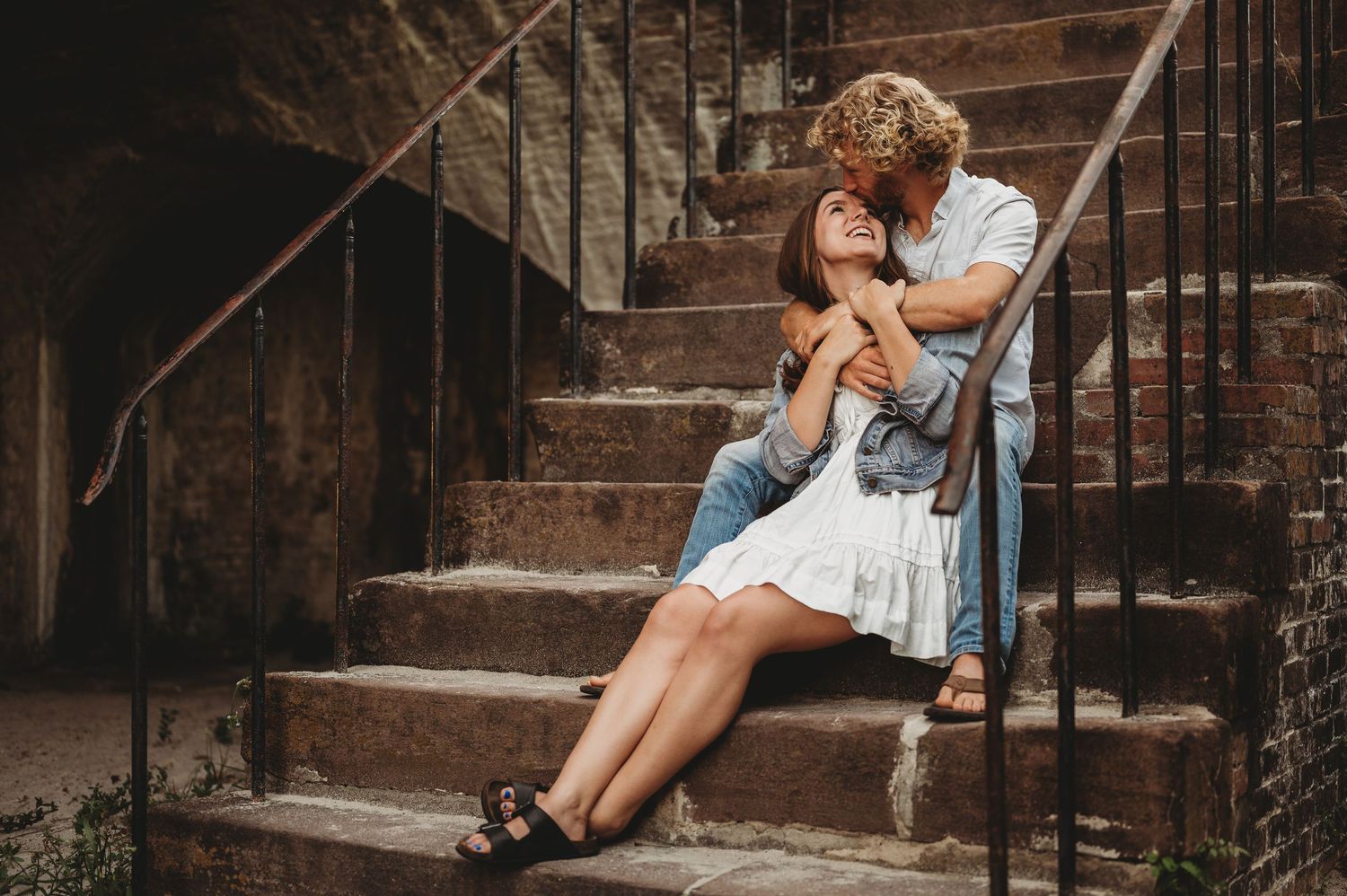 Romantic couple embracing on rustic stone steps, woman in white dress with man kissing her head.