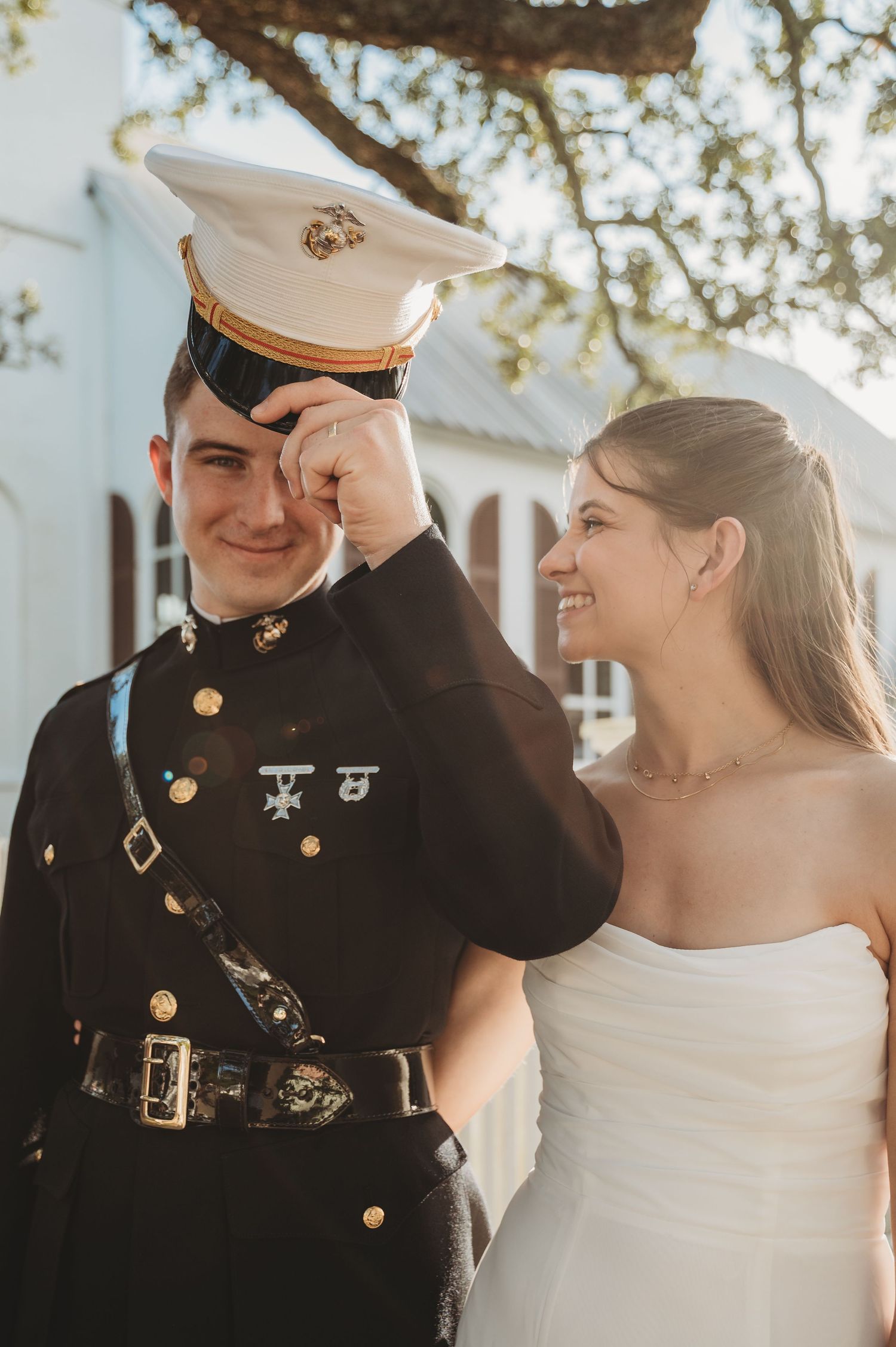 Marine groom in dress uniform wipes tear as smiling bride in white strapless gown looks on outdoors.