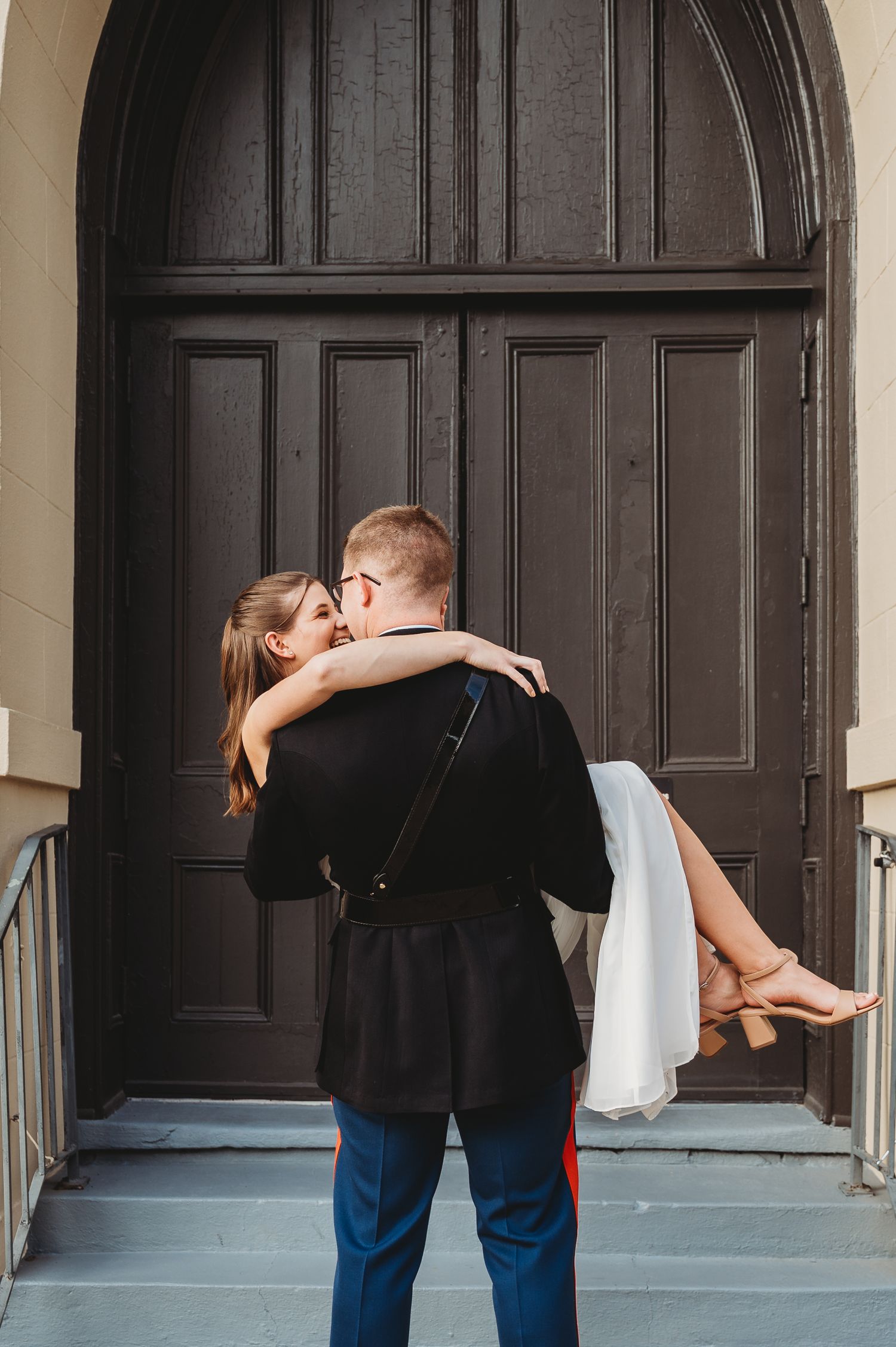 Marine groom carries bride in white gown in front of large arched wooden doors.