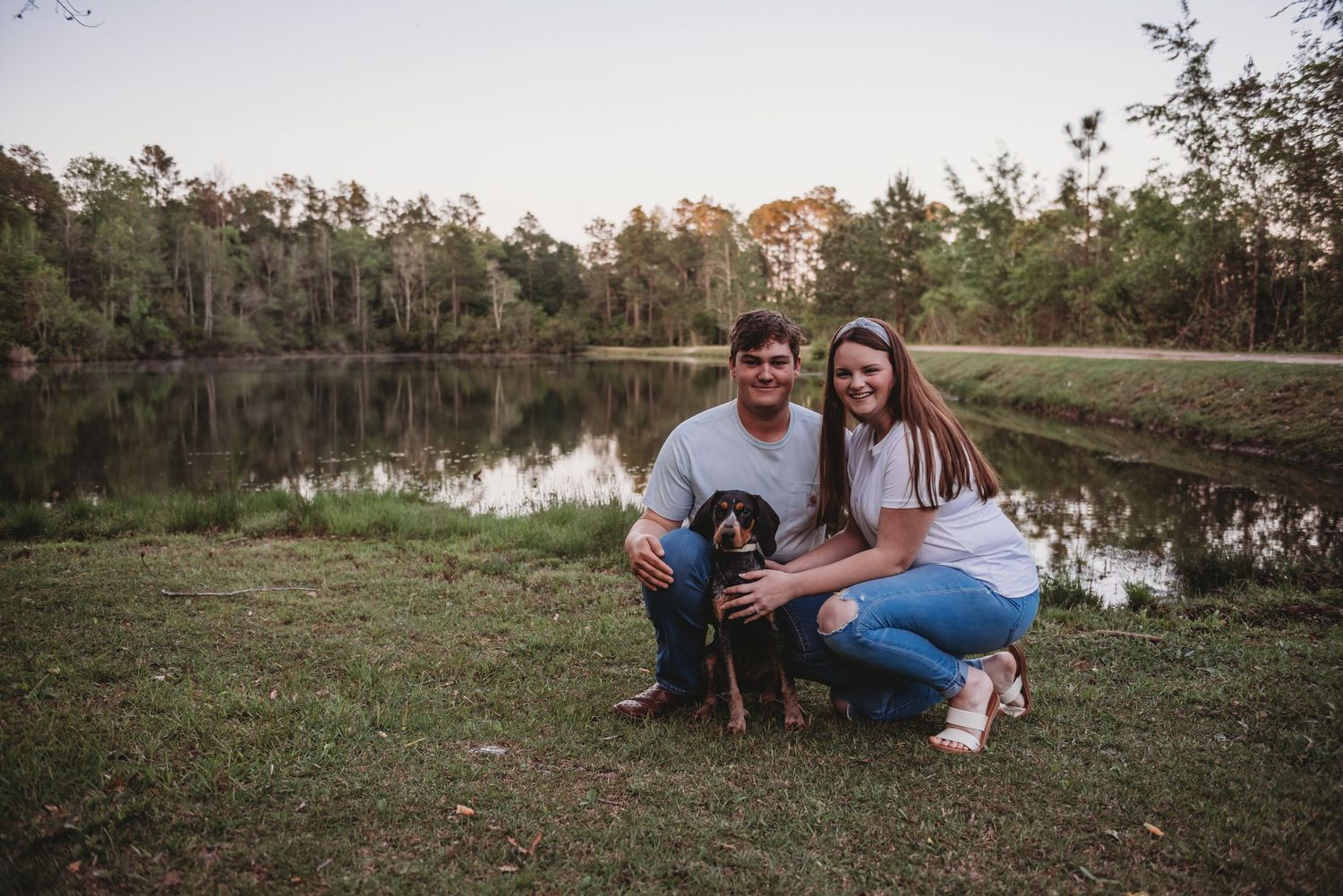 Happy couple kneeling with their black dog beside a tranquil pond at dusk.