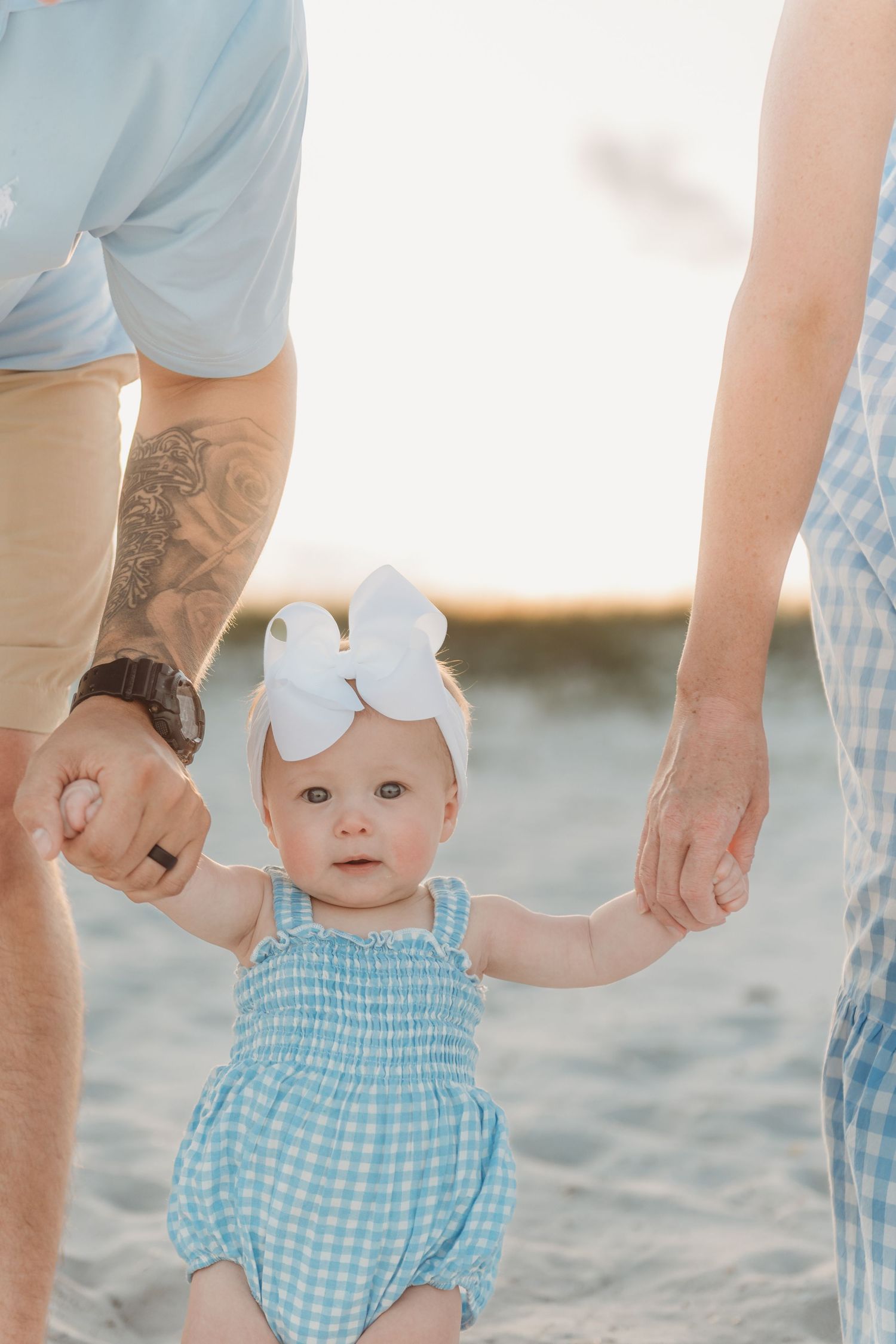 Adorable baby girl in blue gingham romper holding parents hands on the beach.