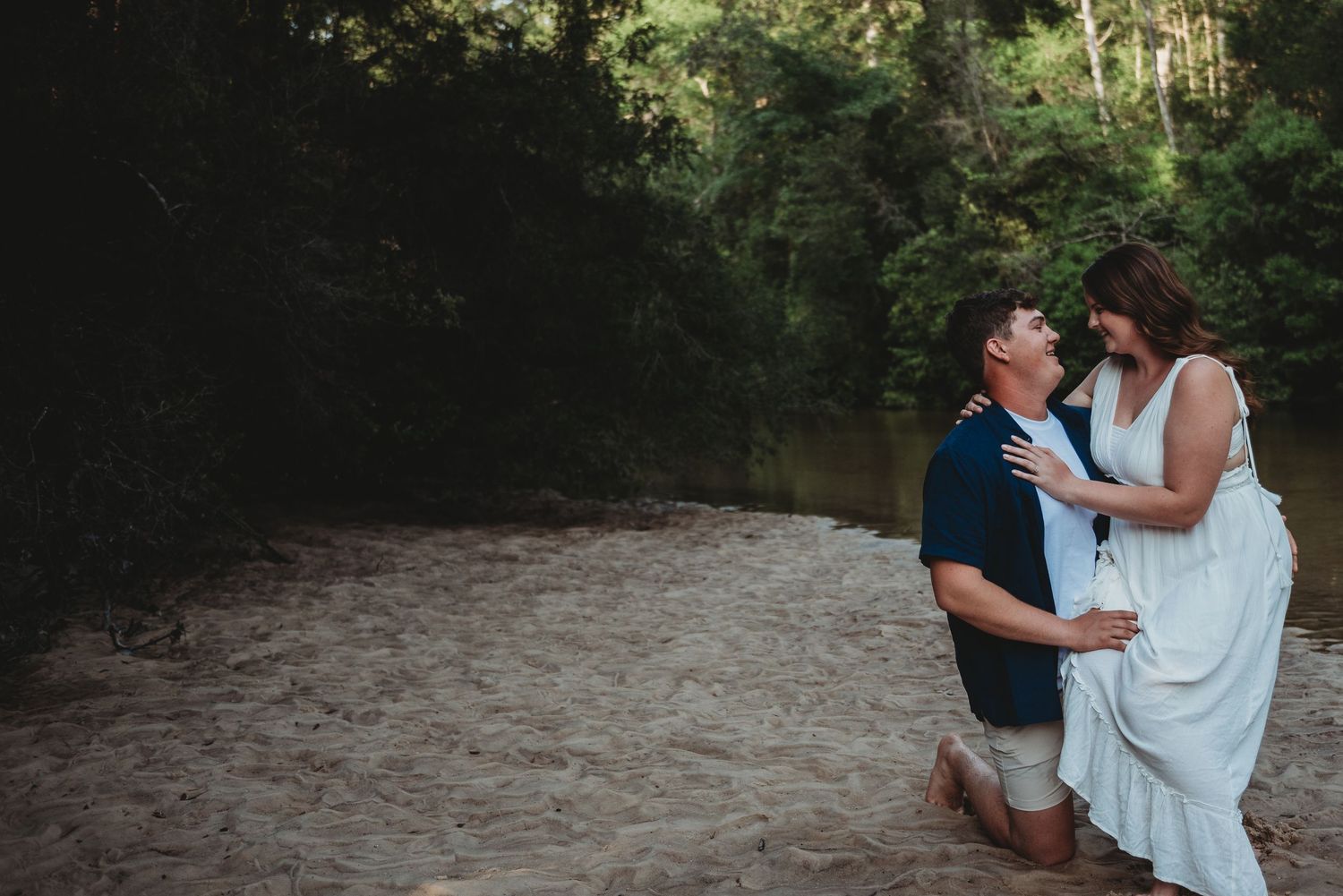 Man kneeling on sandy riverbank proposing to woman surrounded by lush green forest trees.