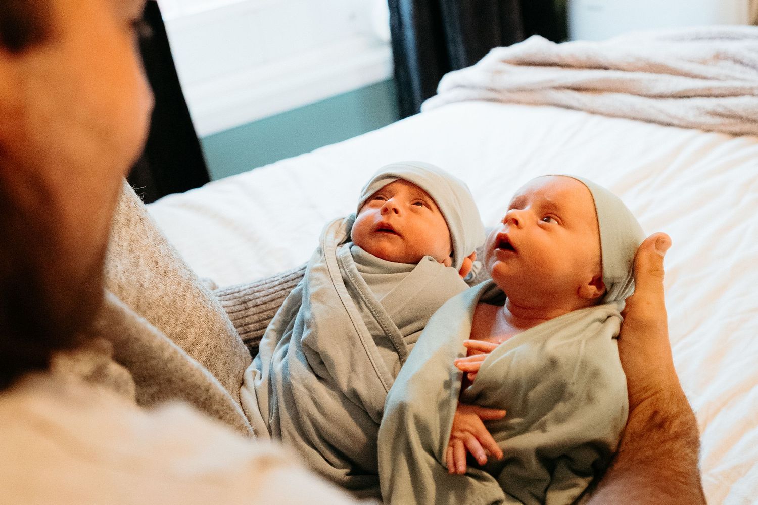 Mother holding two swaddled newborn twin babies in matching light blue wraps and knotted hats.