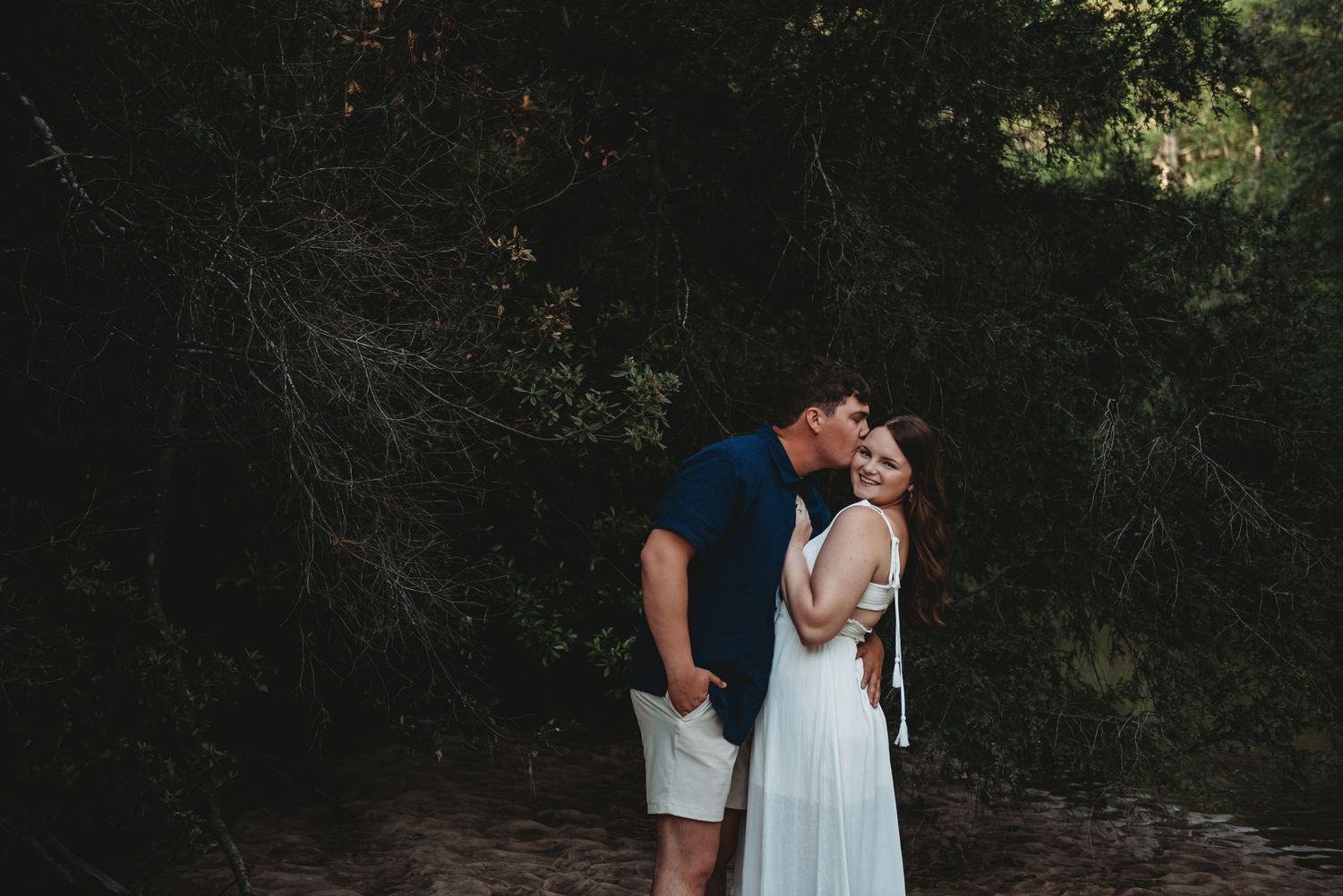 Couple sharing a romantic kiss on a sandy path surrounded by dark green woodland trees.