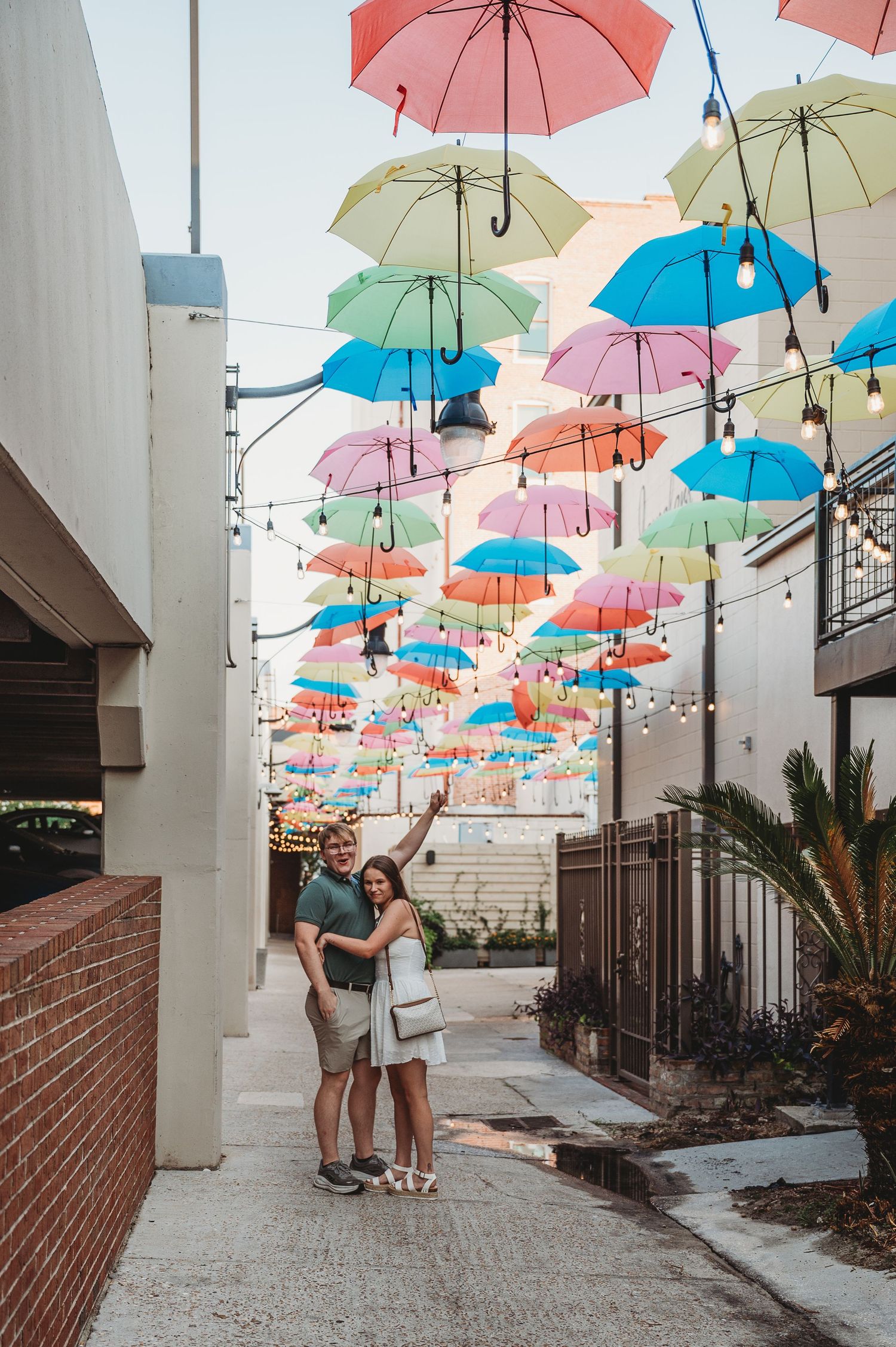 Happy couple pointing up at vibrant colorful umbrellas suspended above a charming outdoor alley.