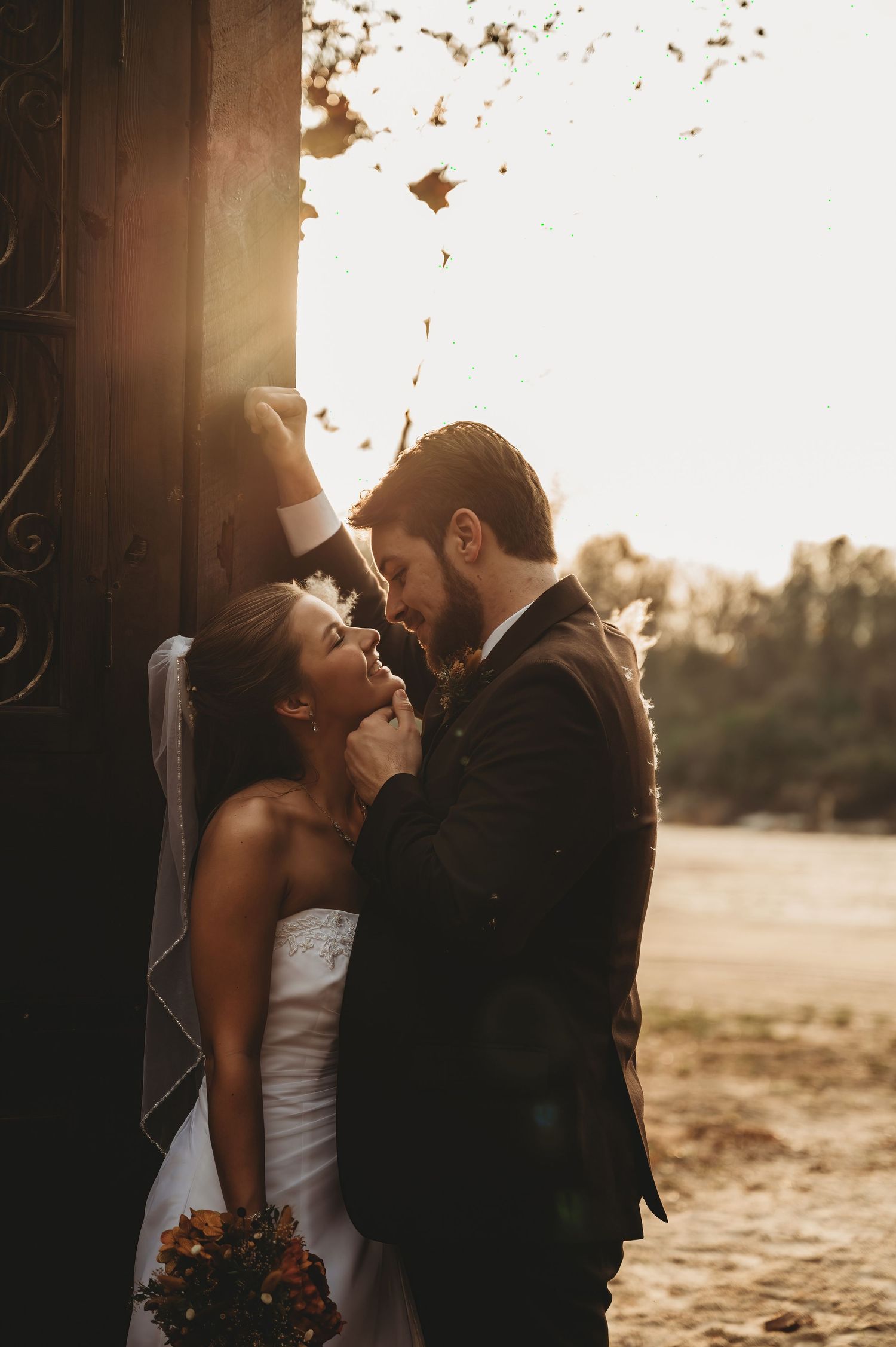 Bride and groom share a romantic moment at golden hour, leaning in close near an ornate door with falling leaves.