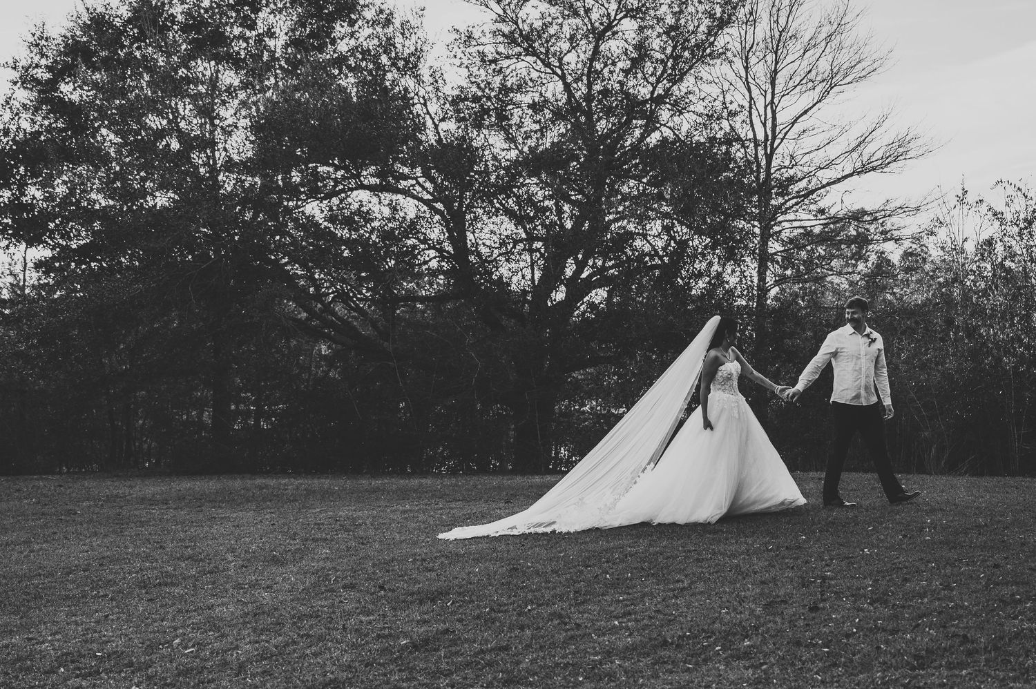 Black and white photo of a bride and groom holding hands walking across a grassy field with trees in the background.