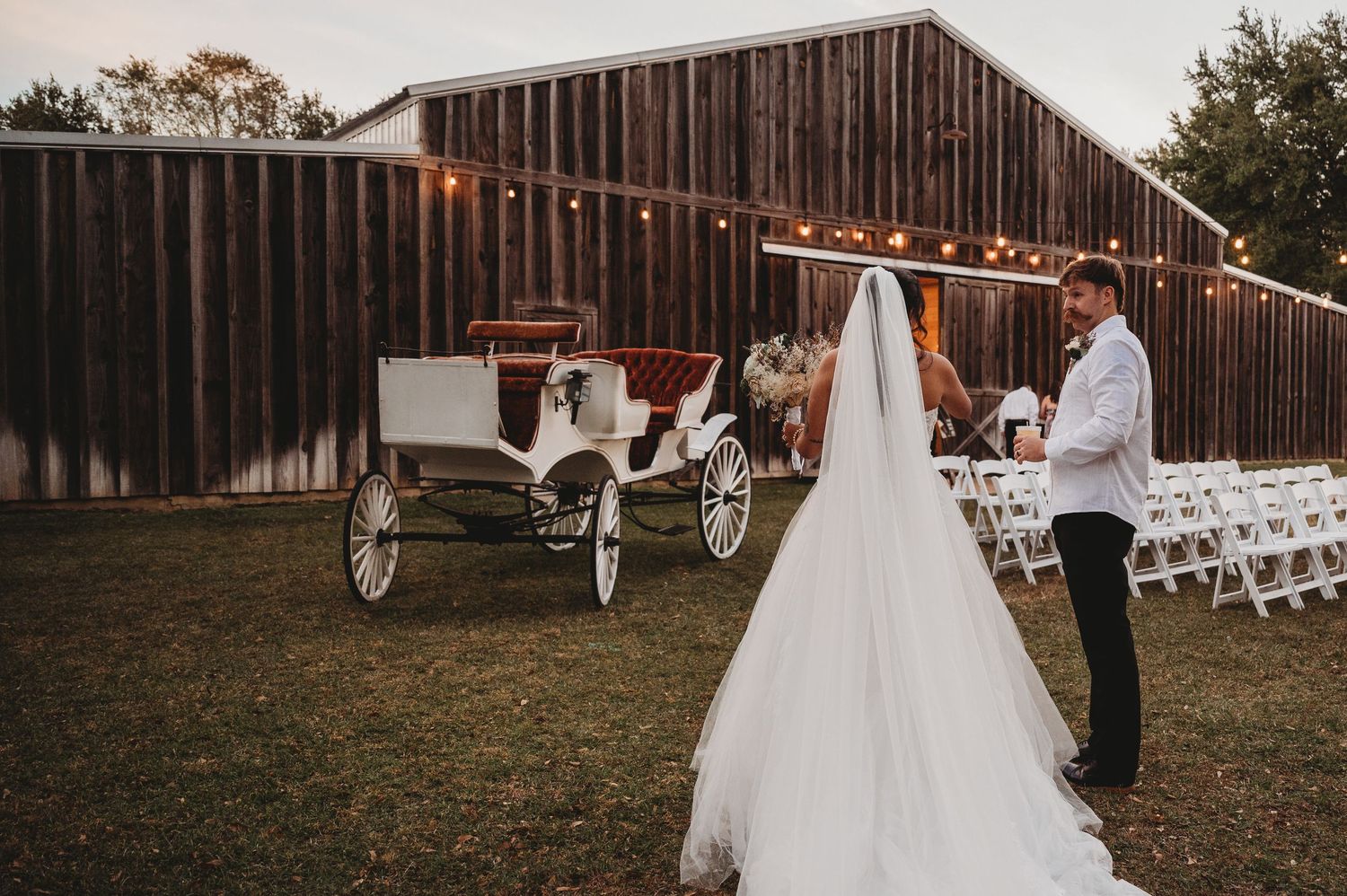 Bride and groom walk toward a white horse-drawn carriage at a rustic barn wedding venue with string lights.
