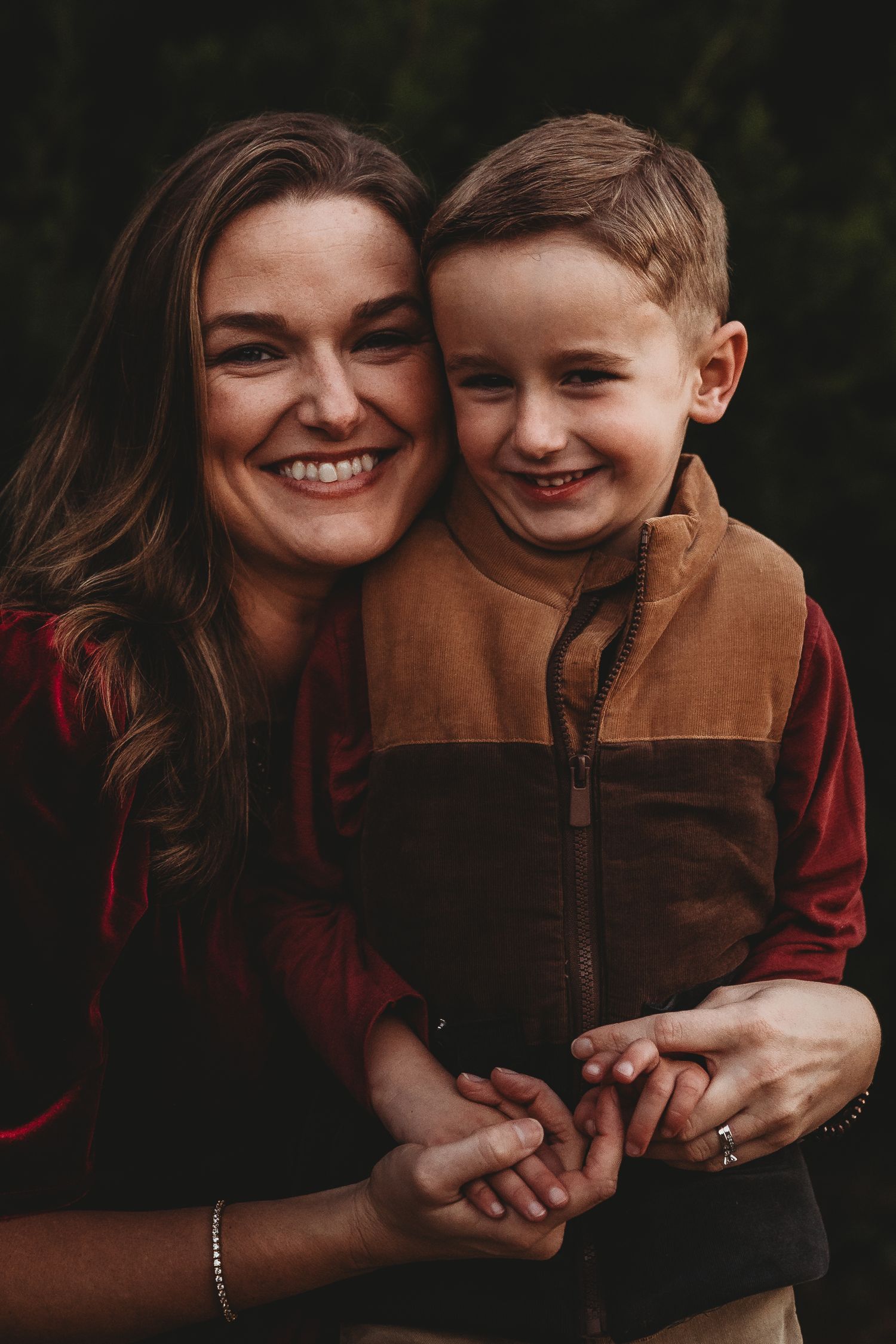 A smiling woman hugs a young boy in brown vest outdoors, both happy in a warm family portrait.