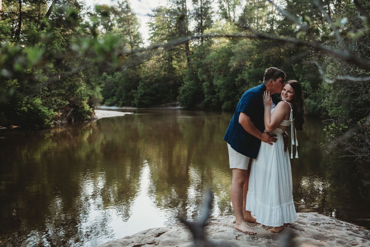 Engaged couple embracing near a peaceful river surrounded by green trees during golden hour.