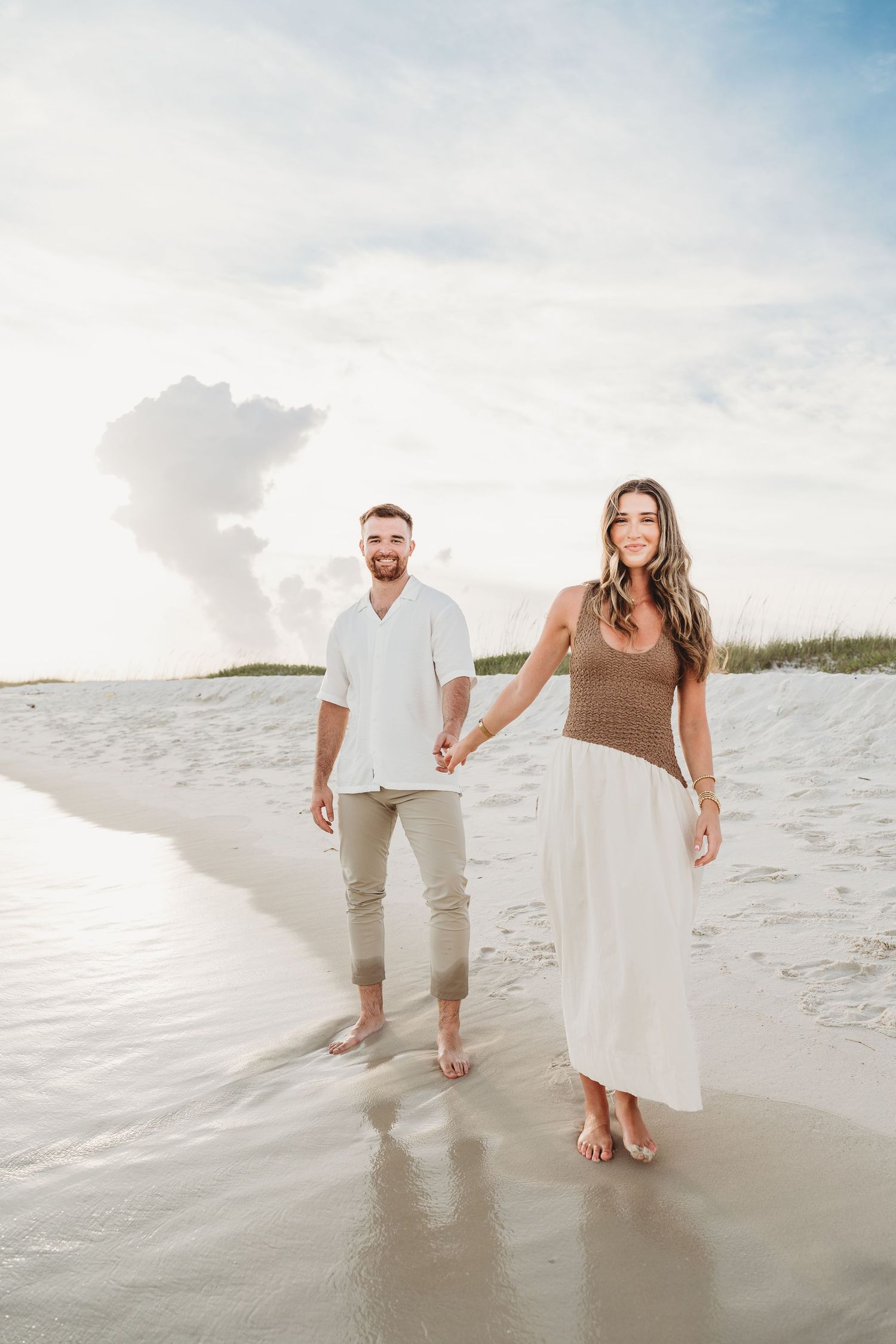 A couple smiles while walking barefoot on a sunlit sandy beach, holding hands together.
