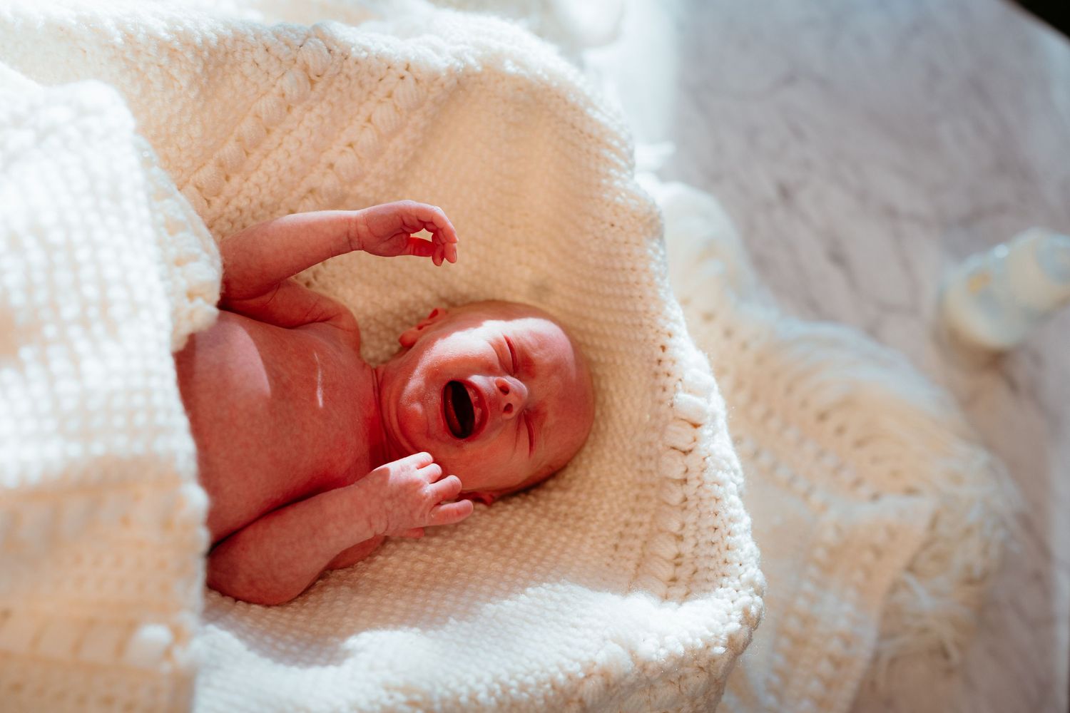 Crying newborn baby lying on a white knit blanket in a wicker bassinet with arms raised.