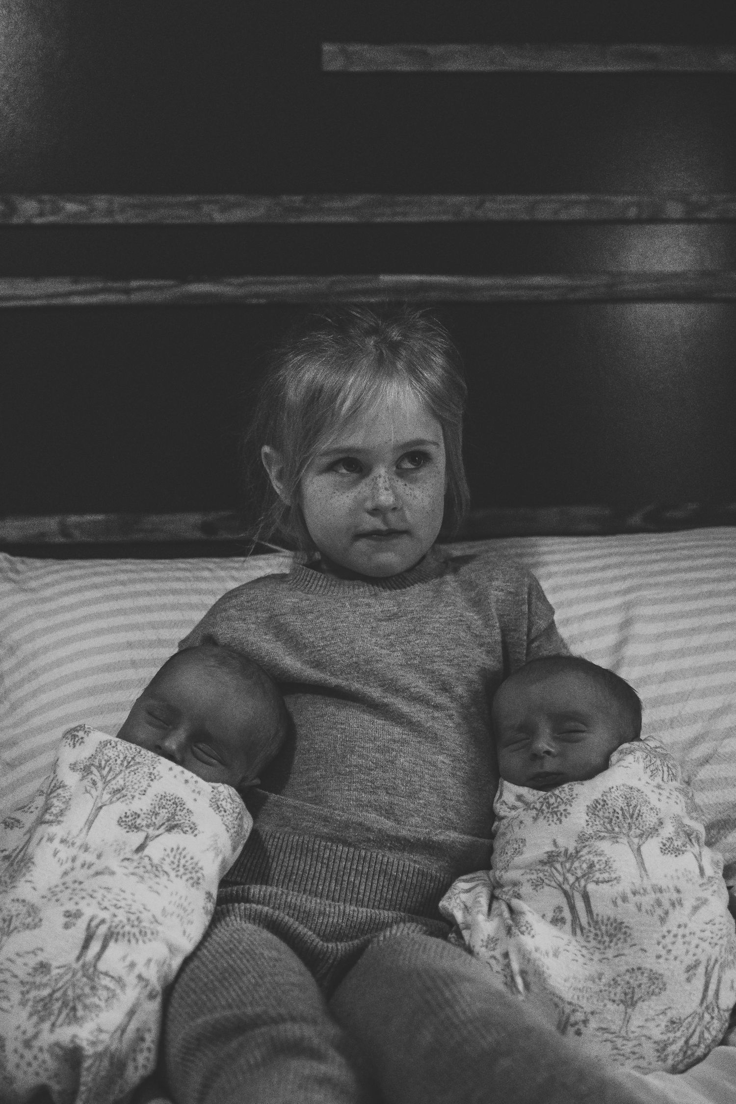 Black and white photo of a young child sitting on a bed holding two sleeping newborn twins.