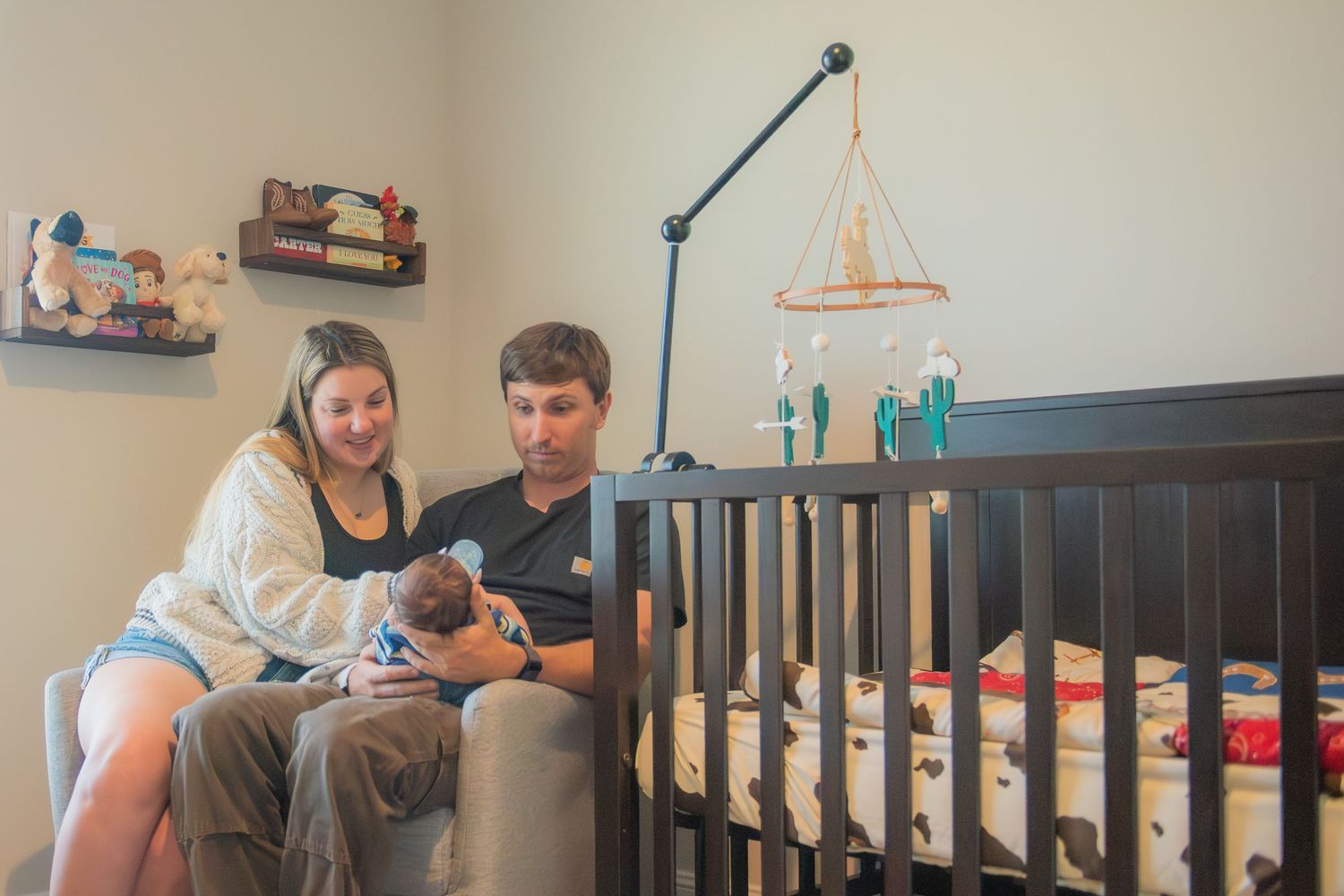 Happy parents sitting together near crib holding their newborn baby in nursery.