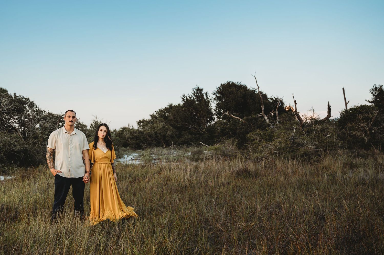 Couple standing in golden meadow at sunset, woman in flowing mustard yellow dress.