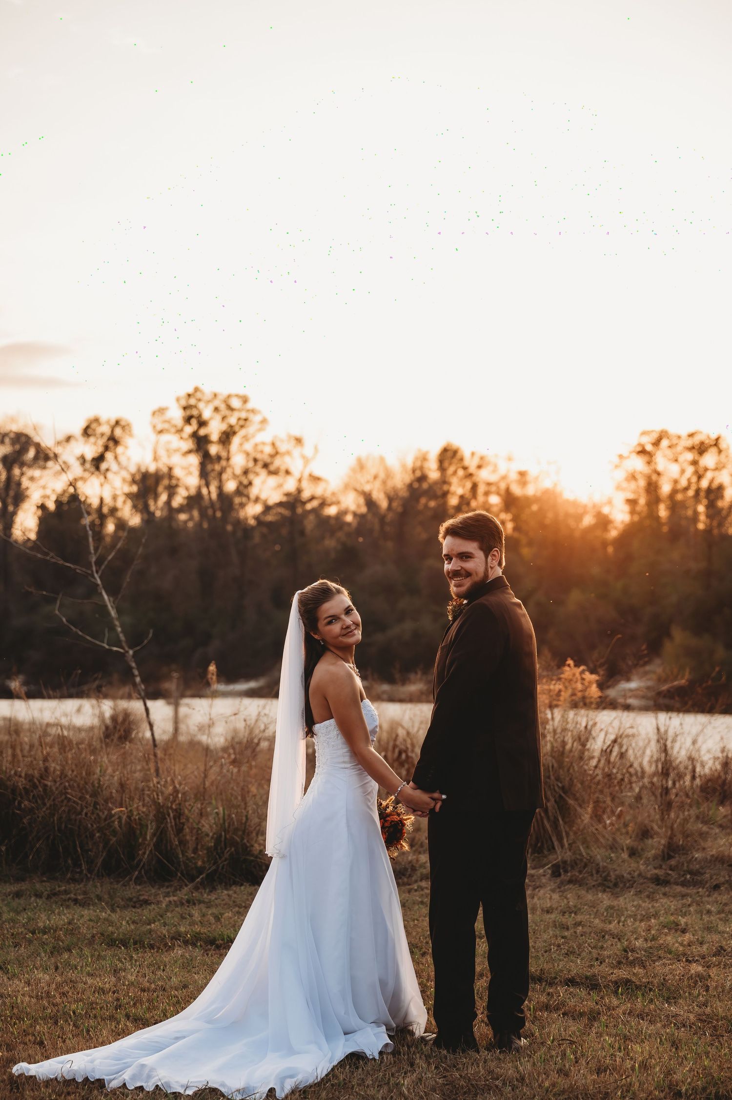 Bride in white gown and groom in black suit hold hands at sunset by a serene lake in an autumn field.
