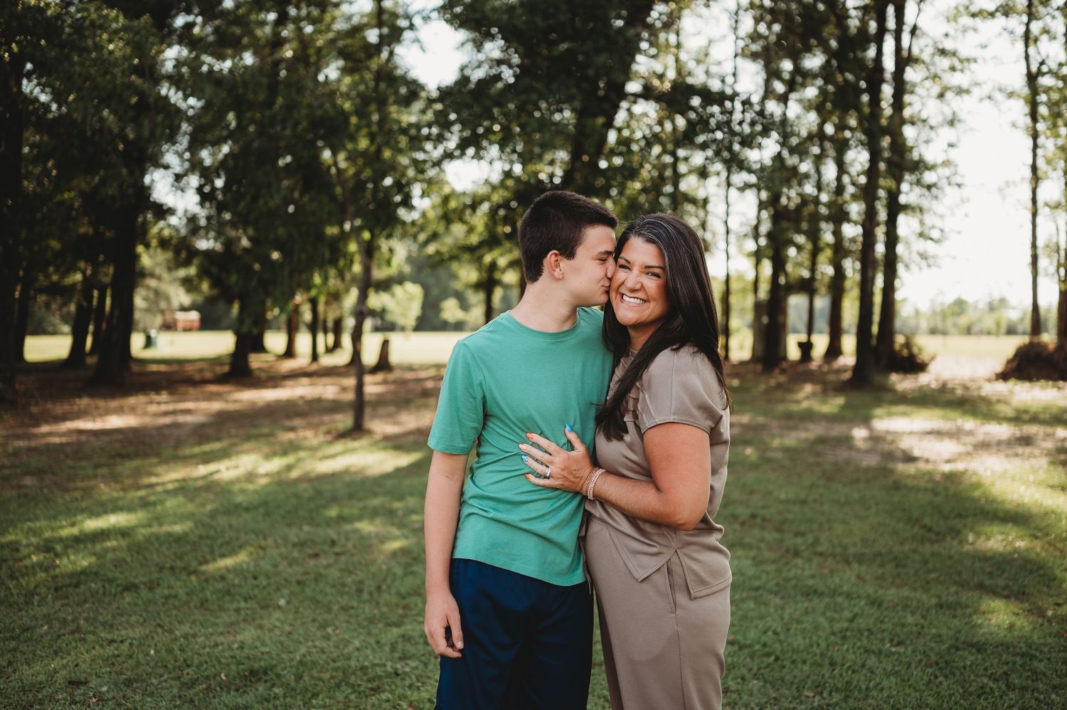 A couple embraces and smiles in a lush green park surrounded by tall trees on a sunny day.