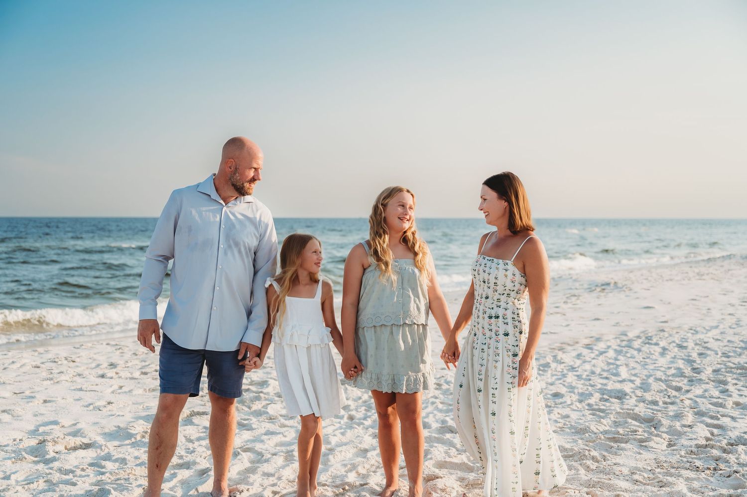 Family of four holding hands and walking on a sunny beach, wearing coordinated white and blue outfits.