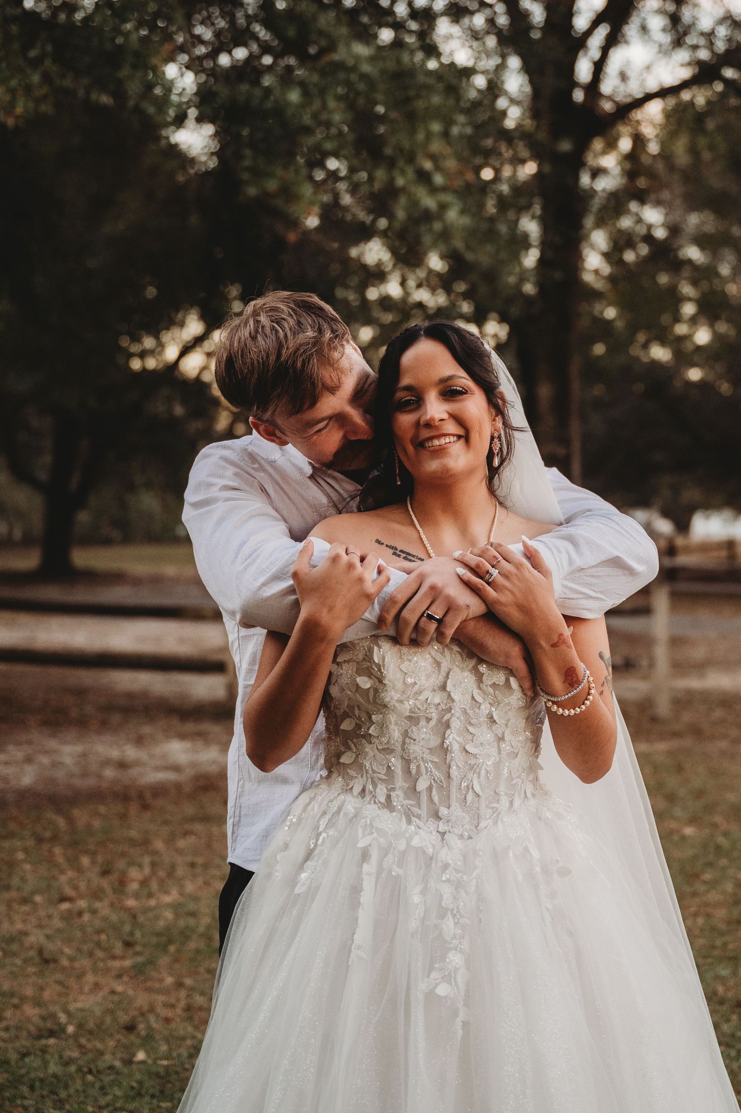 A groom in a white shirt embraces his smiling bride in a lace wedding gown outdoors among trees.