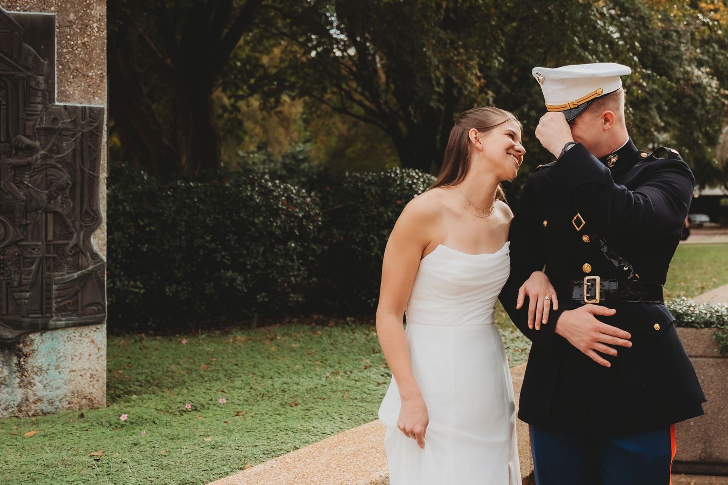 A bride in a white gown laughs beside a Marine in dress uniform adjusting his cap outdoors.