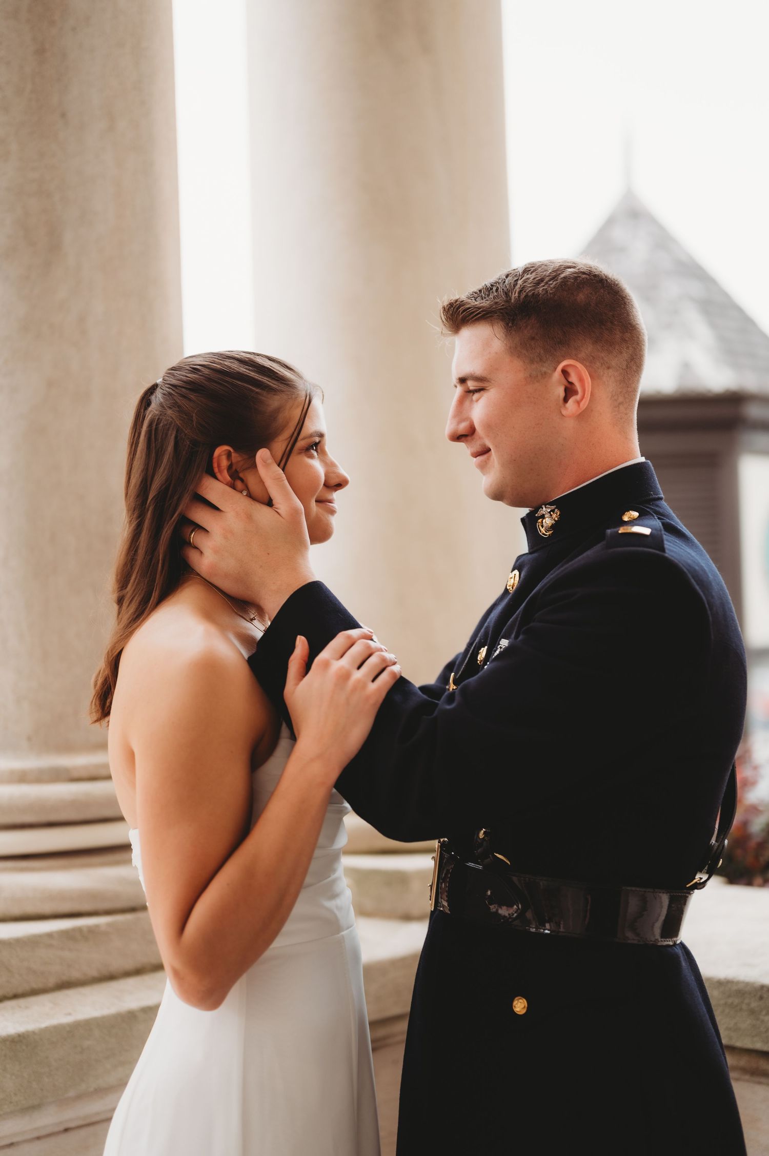 Military groom in dress uniform tenderly holds bride's face during romantic outdoor wedding portrait by stone columns.