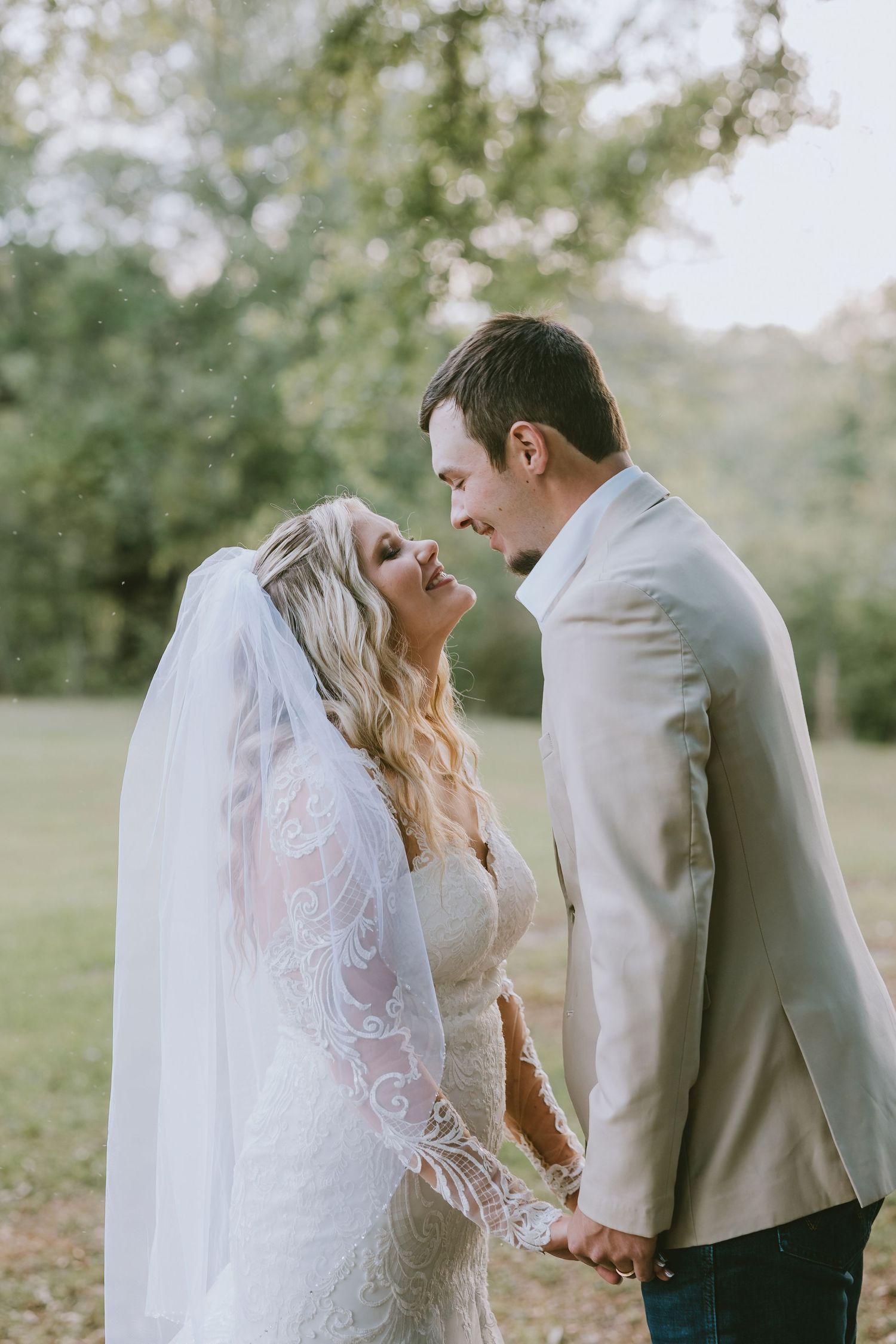 Bride in lace gown and veil gazes up at groom in tan suit as they hold hands outdoors at golden hour.