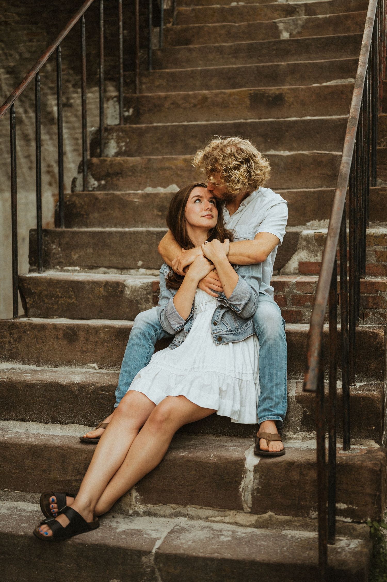 Couple sitting on stone steps, man with curly hair embracing woman in white dress, romantic outdoor portrait.