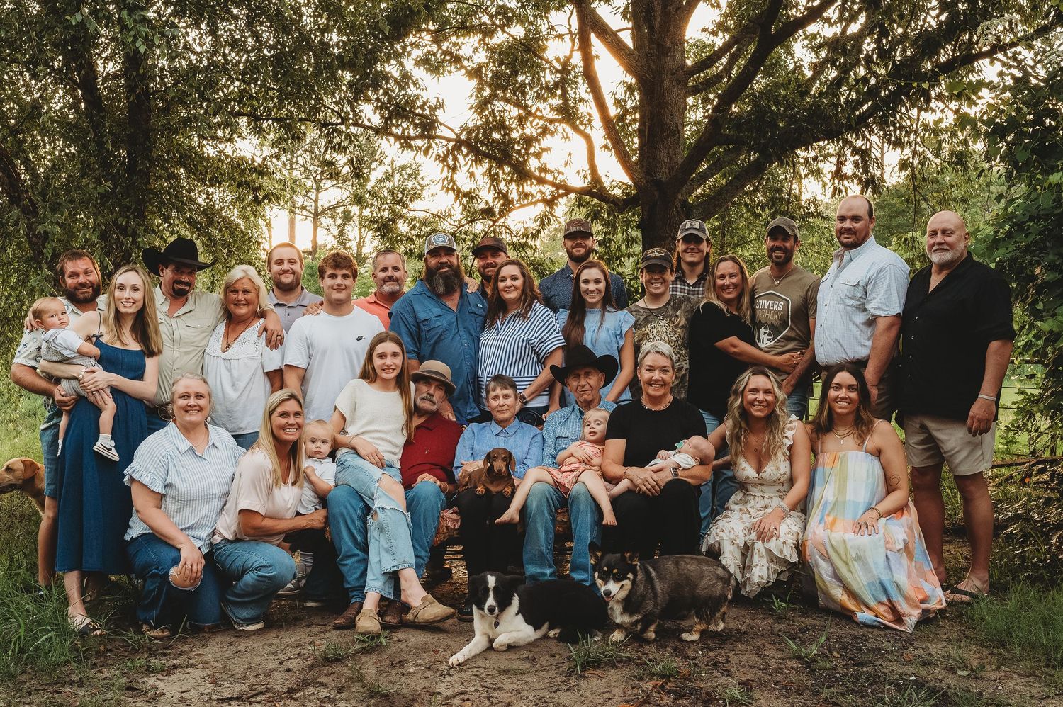 Large family group portrait outdoors under trees, multi-generational gathering with adults, children, and pets on a farm.
