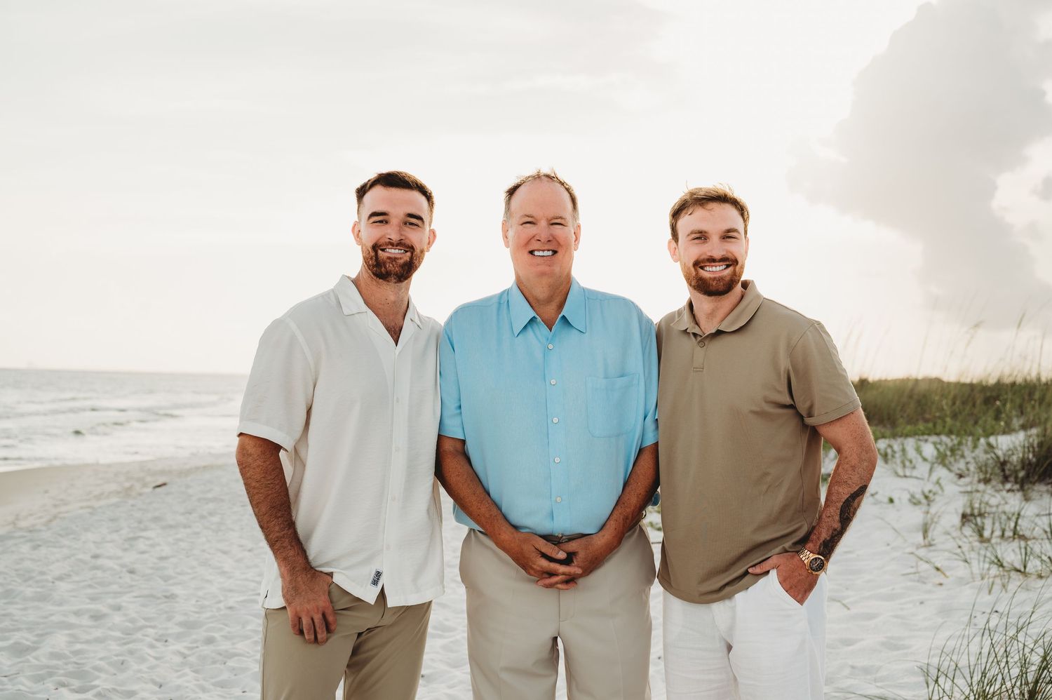 Three smiling men pose together on a beach with sand dunes and ocean in the background.