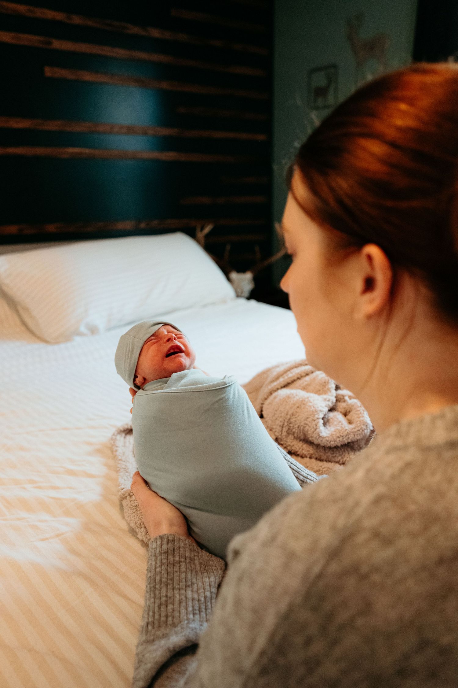 Red-haired mother holding her swaddled newborn baby in a light blue wrap on a white bed.