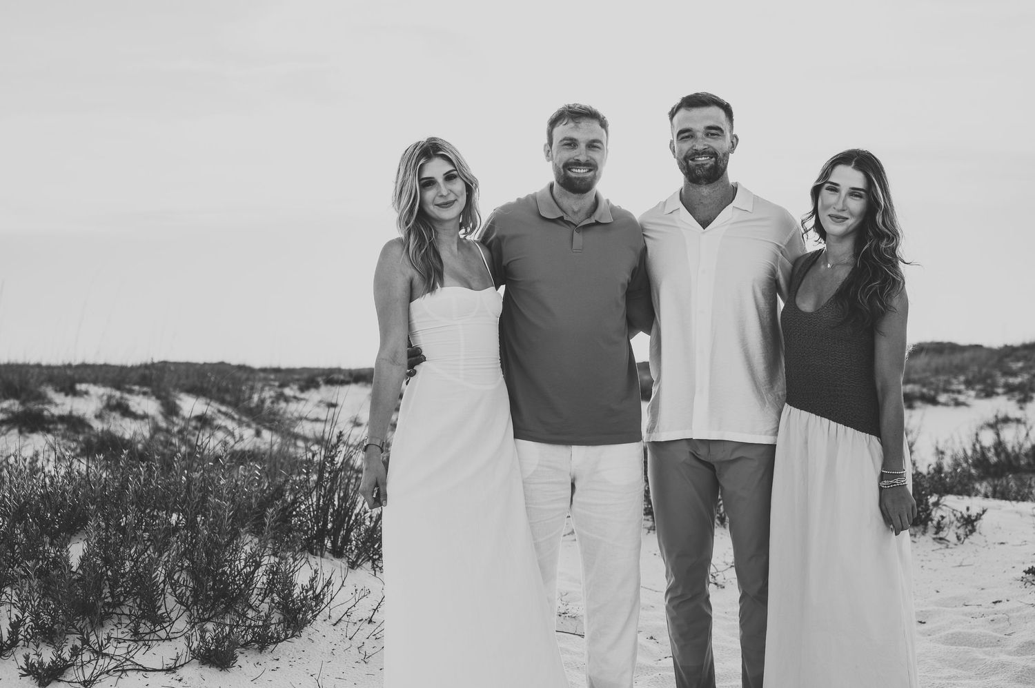 Black and white photo of four young adults smiling together on a sandy beach with dunes behind them.