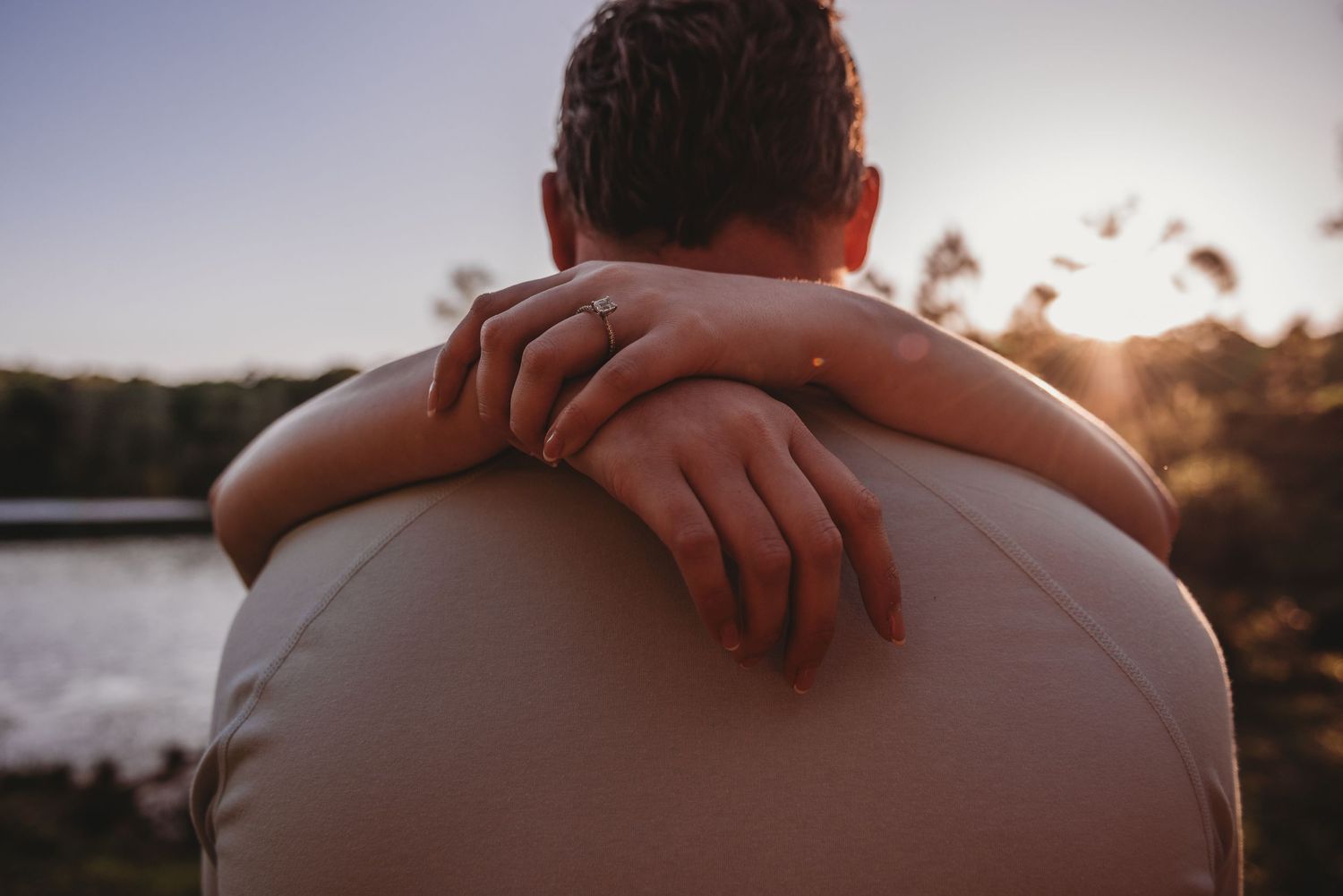 Close-up of woman's hands with engagement ring wrapped around man's shoulders at sunset by water.